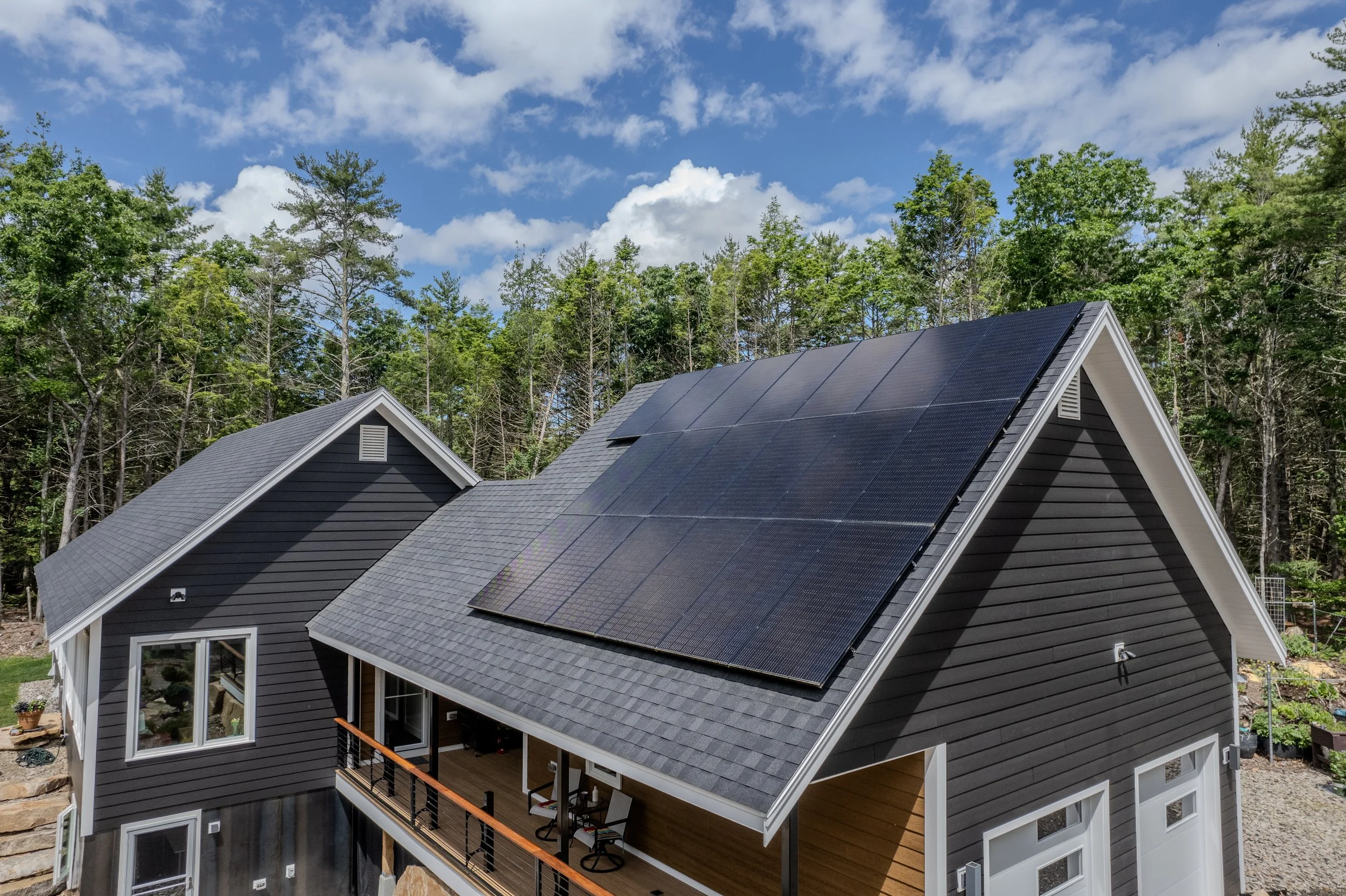 A house with dark gray siding and a large array of solar panels installed on its roof, surrounded by trees under a partly cloudy sky.