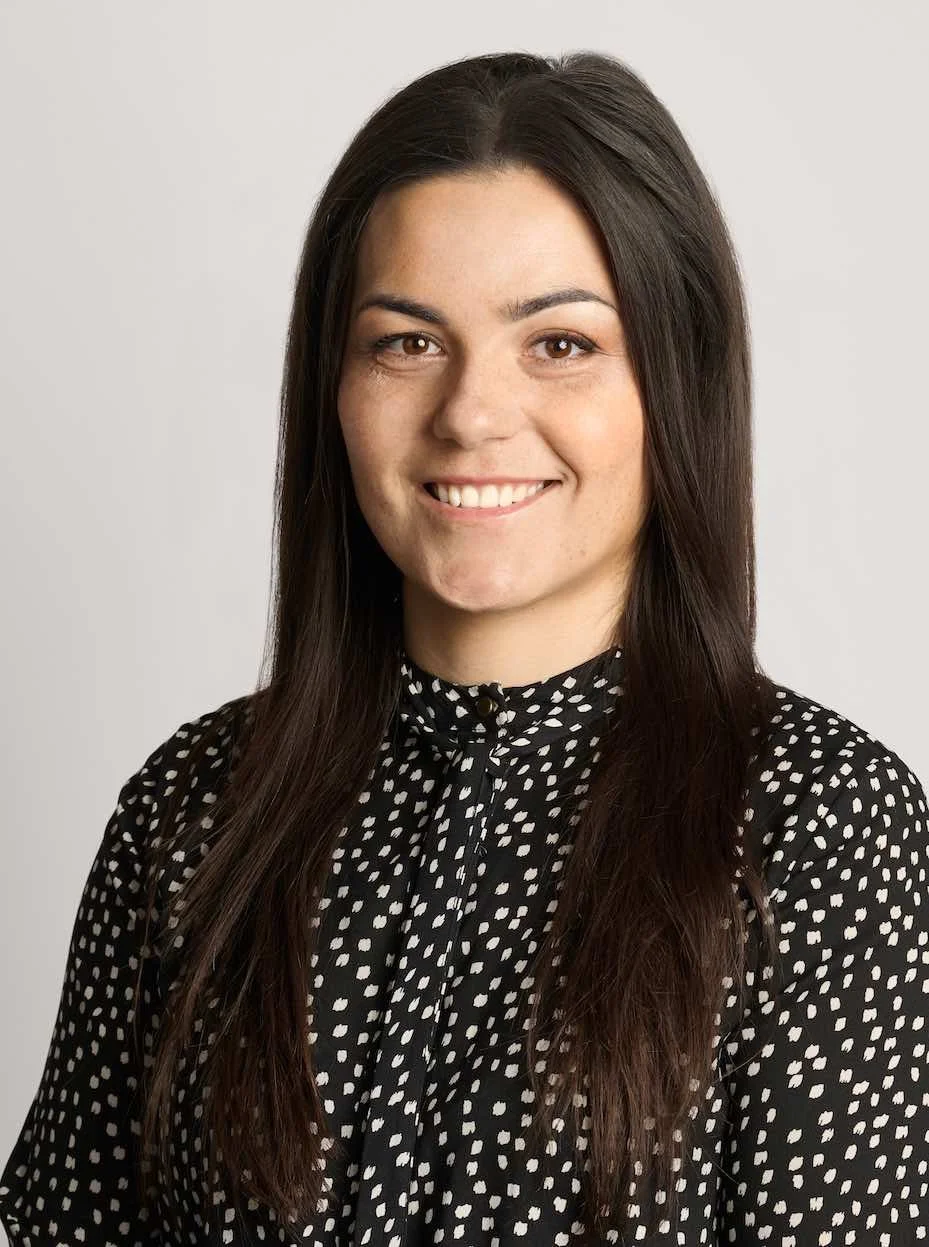 A woman with long dark hair wearing a black and white polka dot blouse, smiling against a plain background.