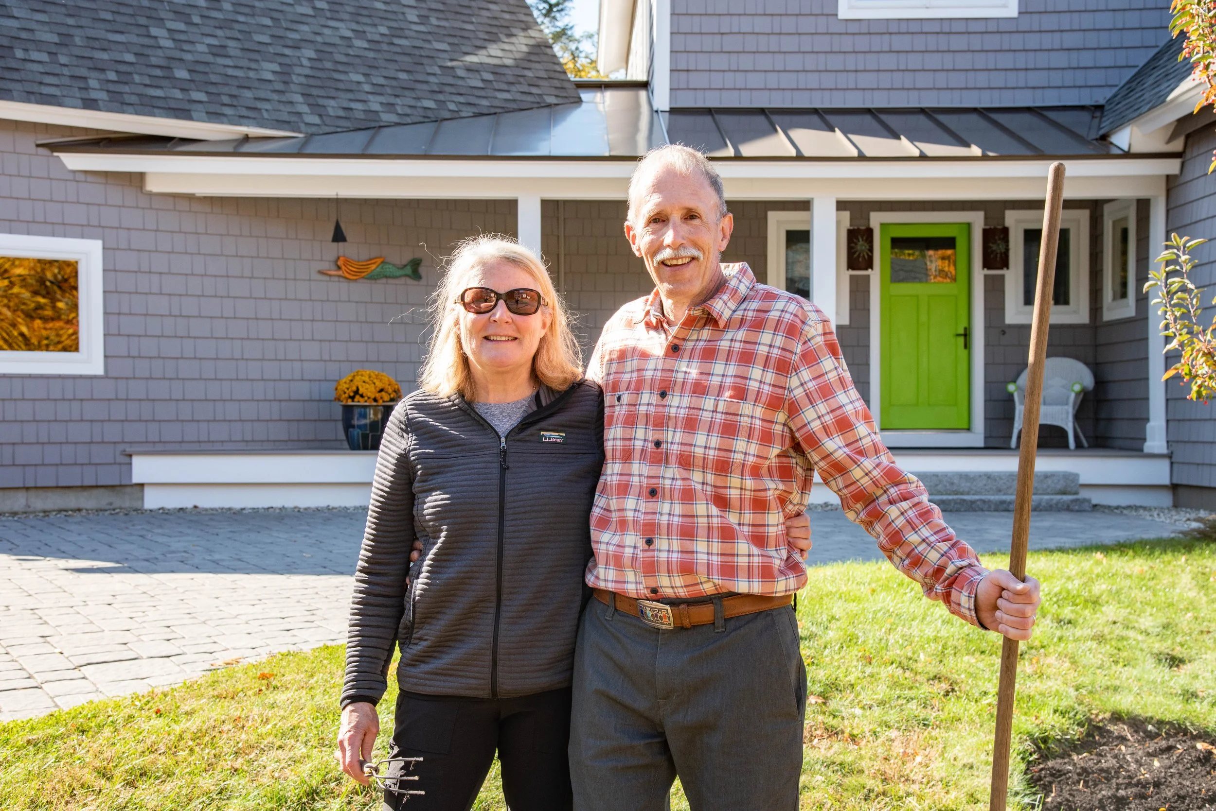 A happy elderly couple standing outdoors in front of a gray house with a bright green door, smiling and posing for a photo on a sunny day.