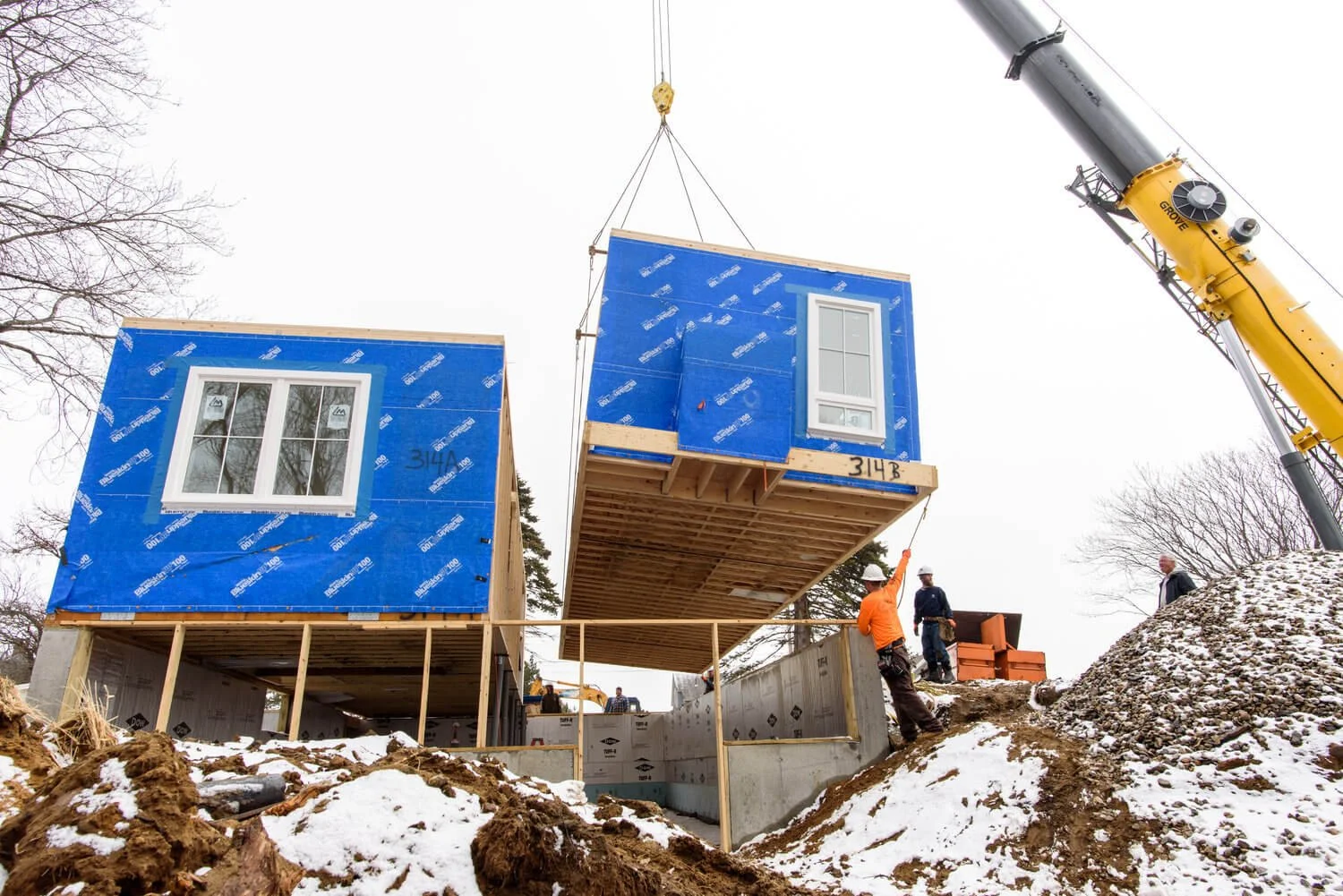 Construction workers guide a prefabricated blue modular home into place with a crane on a snowy day.