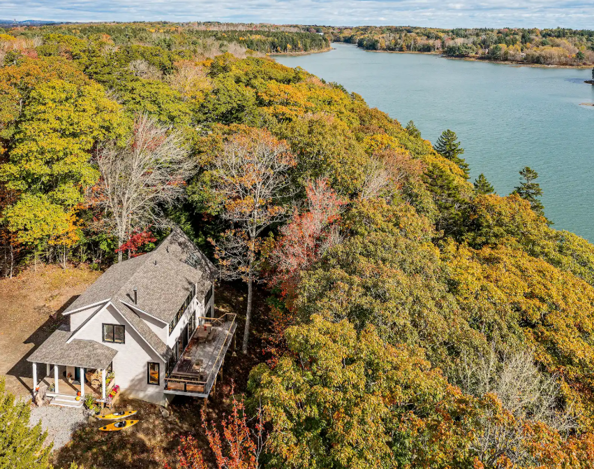 A house with a porch and deck next to a forested area with colorful autumn foliage, near a large wide river.