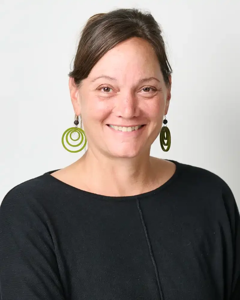 Smiling woman with short brown hair wearing green hoop earrings and a black top against a plain white background.