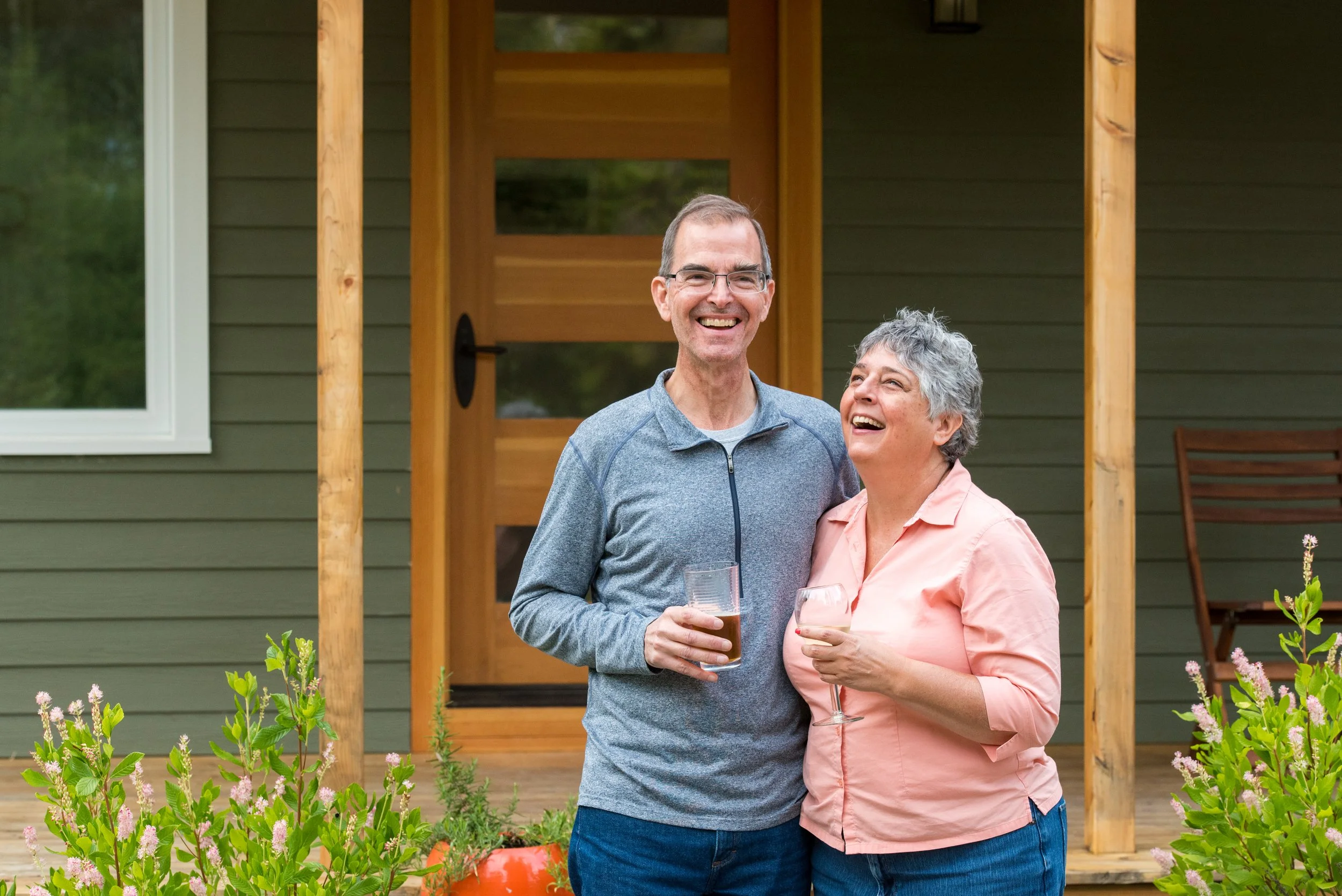 A smiling older man and woman standing outside a house, holding drinks, enjoying a conversation. The man has short brown hair and glasses, wearing a gray zip-up jacket, and the woman has short gray hair, wearing a light pink shirt. There are flowering plants in the foreground and a wooden deck with a door and window behind them.