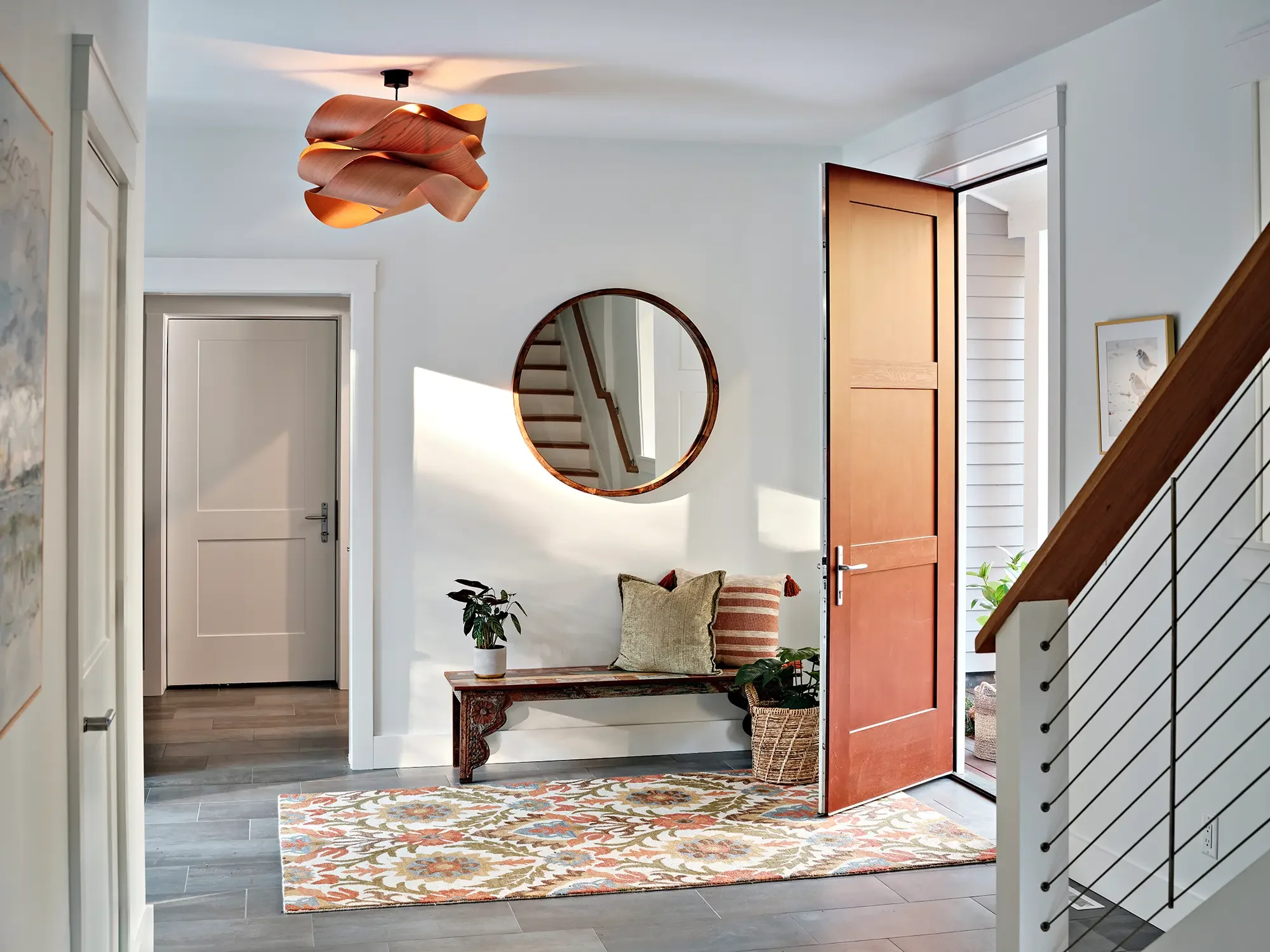 Bright modern entryway with wooden front door, round mirror, bench with pillows, and patterned rug.