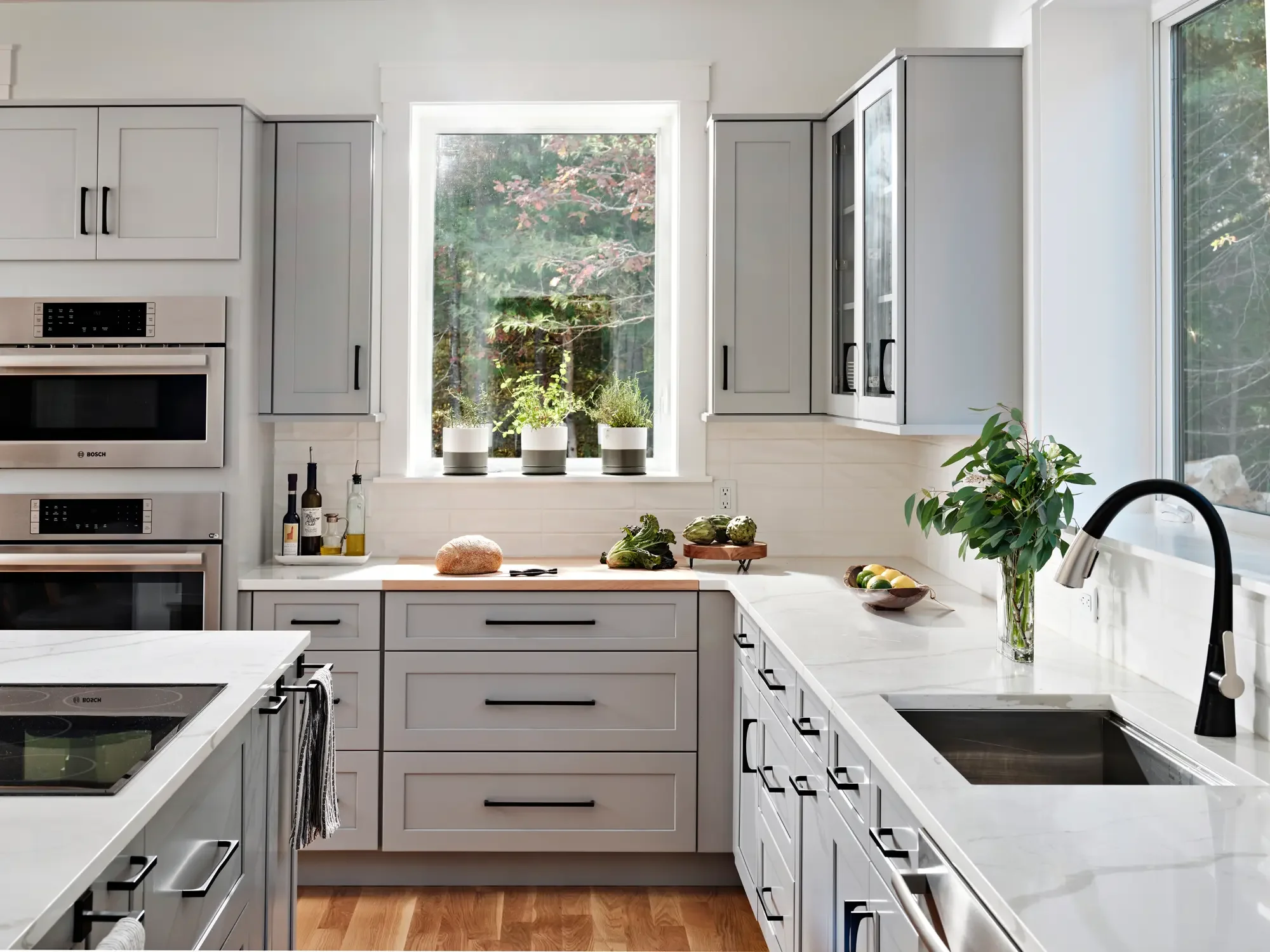 Modern white kitchen with gray cabinets, white countertops, stainless steel appliances, and a large window with greenery outside, decorated with potted plants and fresh vegetables.