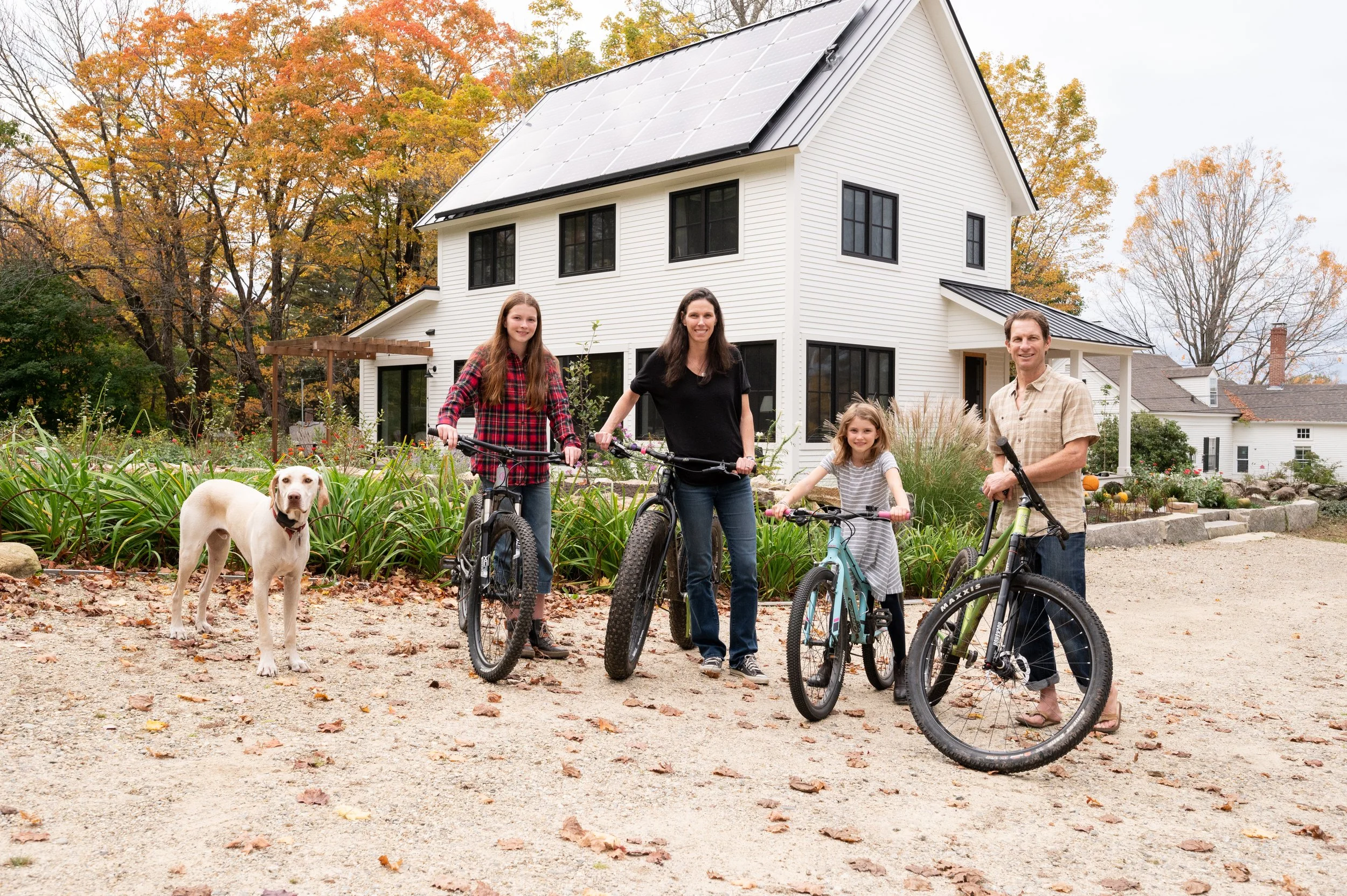 A family of four, with two girls, a man, and a woman, along with a dog, standing with bicycles in front of a white two-story house surrounded by autumn trees and landscaping.
