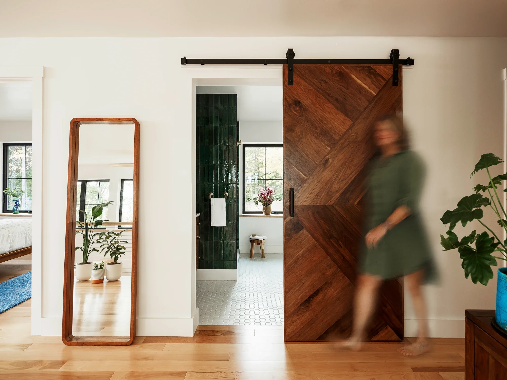 A blurred woman walking past a wooden sliding barn door in a modern home interior, with plants and a full-length mirror visible.