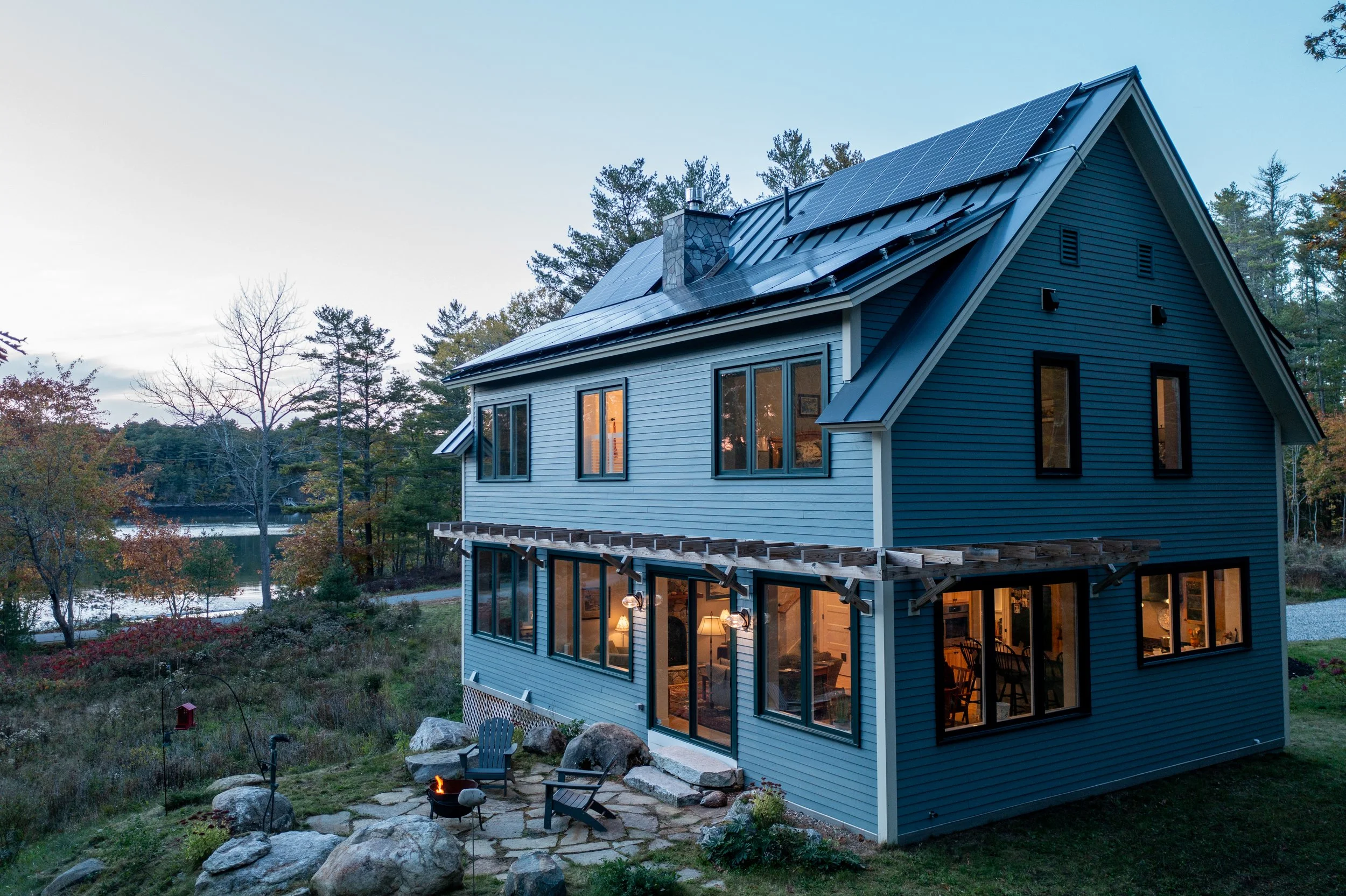 A two-story blue house with large windows and solar panels on the roof, situated in a wooded area near a lake, during dusk.