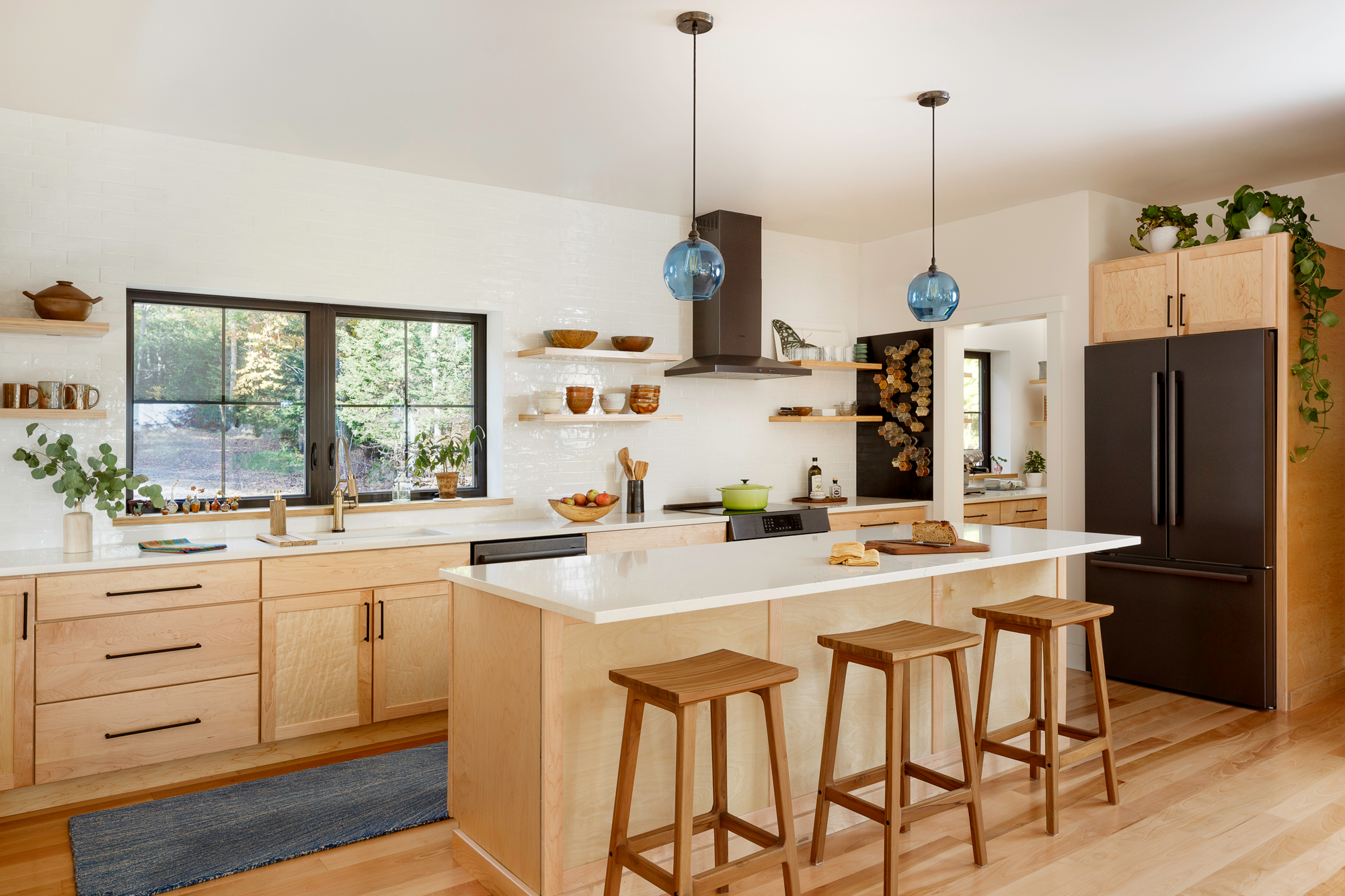 Modern kitchen with white countertops, light wood cabinets, black refrigerator, stovetop, window with greenery outside, decorative shelves with bowls and cups, wooden barstools, pendant lights, and various kitchen items.