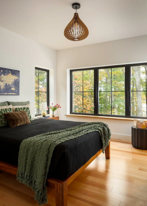 Bedroom with black bed, green throw blanket, pillows, wooden nightstand with pink flowers, large windows with trees outside, white walls, hanging wicker light fixture, and wooden flooring.