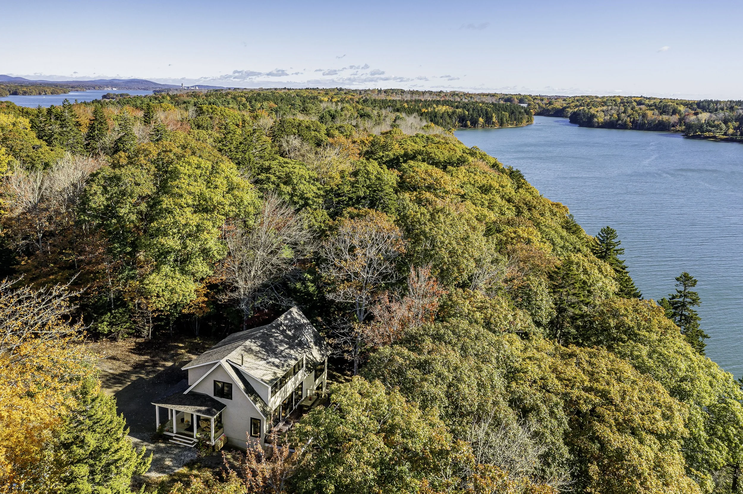 Aerial view of a house surrounded by trees with fall foliage near a large body of water.