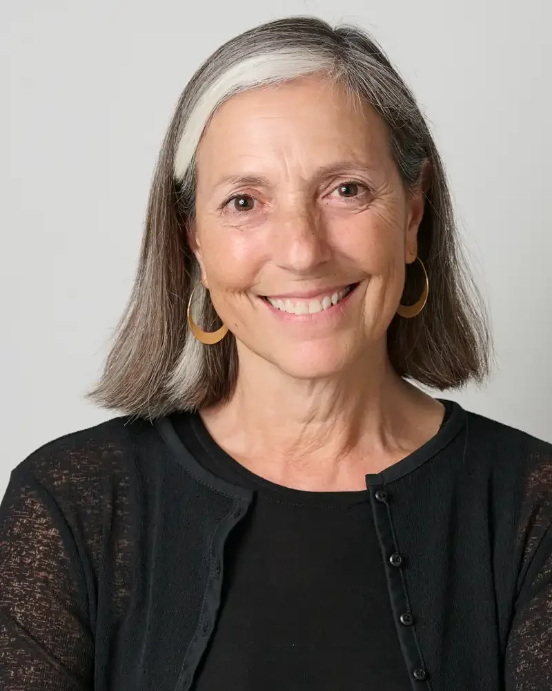 A smiling middle-aged woman with shoulder-length brown and gray hair and hoop earrings, wearing a black top with sheer sleeves and a black cardigan, against a plain light gray background.