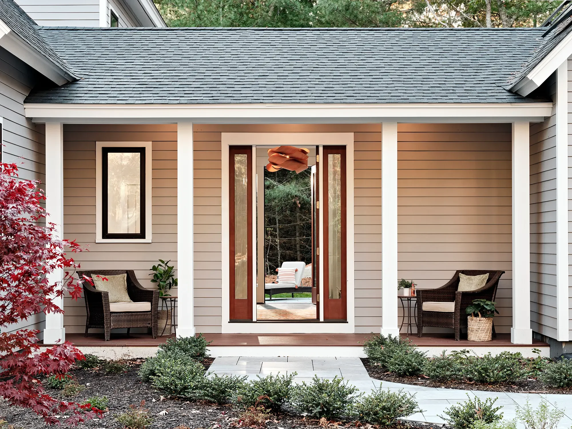 Front porch of a house with white columns, wicker seating on both sides, and open glass double front doors leading to a wooded backyard patio.