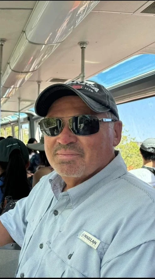 A man wearing black sunglasses, a black cap, and a light blue button-up shirt with a name tag that reads '6 MACEJAN,' sitting inside a bus with other passengers and a blue sky visible outside the window.