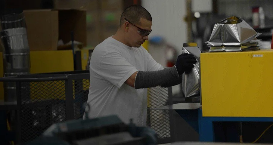 A man wearing safety glasses, black gloves, and a white T-shirt working with a piece of reflective material in an industrial or manufacturing setting.