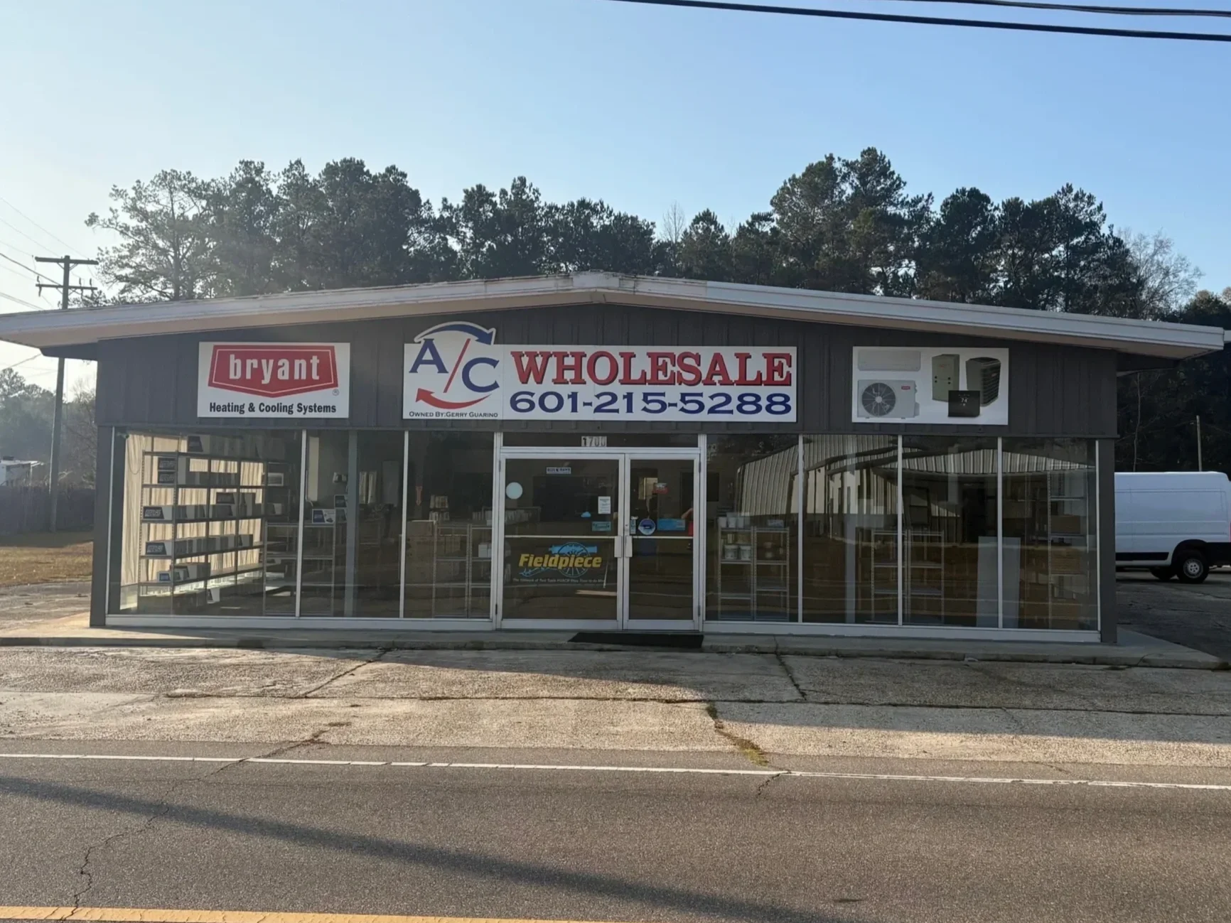 Front view of a small business building with signs for Bryant Heating & Cooling Systems and A C Wholesale, with a phone number 601-215-5288, located on a street with a white van parked to the right and a parking lot.