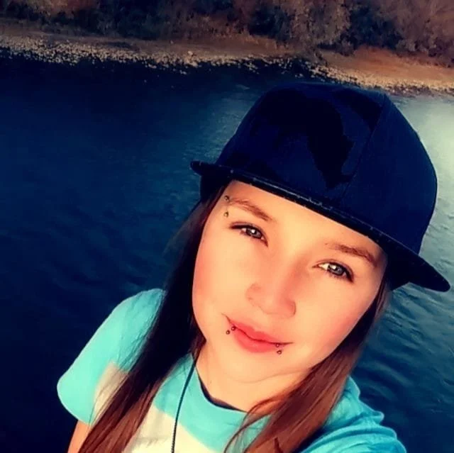 Young woman with piercings wearing a black baseball cap and a light blue shirt, standing near a body of water with a forested shoreline in the background.