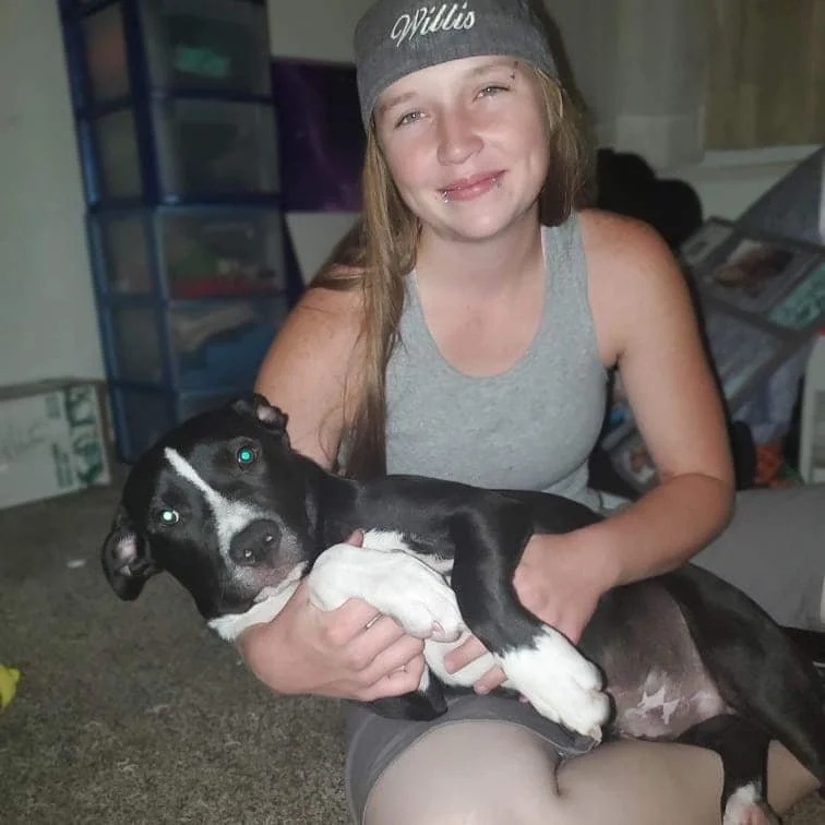A young woman with long hair, wearing a gray beanie and sleeveless shirt, smiling and holding a black and white puppy indoors.