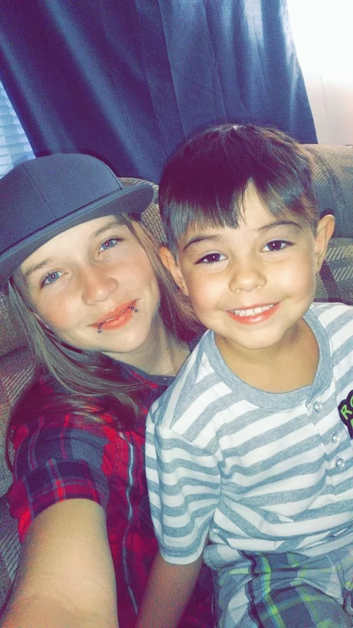 A girl with long hair wearing a dark gray hat and a red plaid shirt sitting on a couch with a young boy in a gray and white striped shirt smiling.