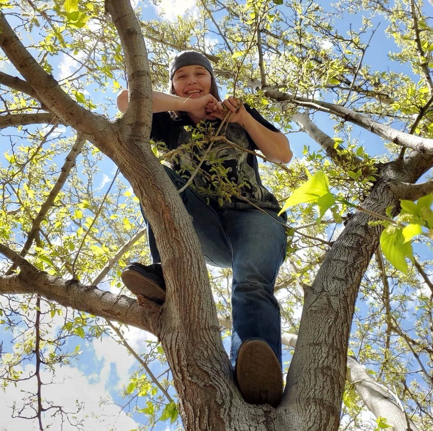 A person sitting in a tree, smiling, wearing a bandana, with green leaves and a blue sky in the background.