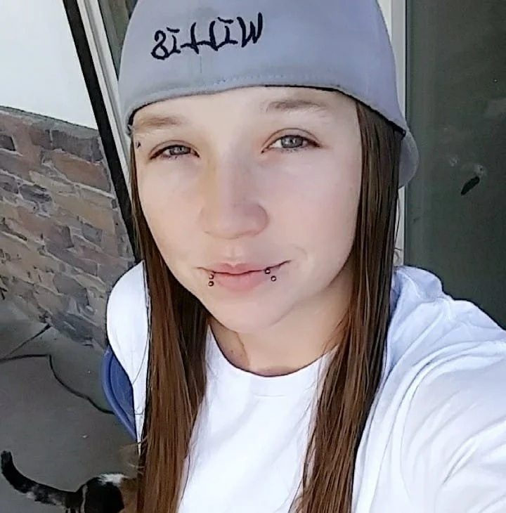A young woman with long brown hair, wearing a gray cap with writing on it, taking a selfie outside near a brick wall and glass door, with a black and white cat partially visible at the bottom of the photo.