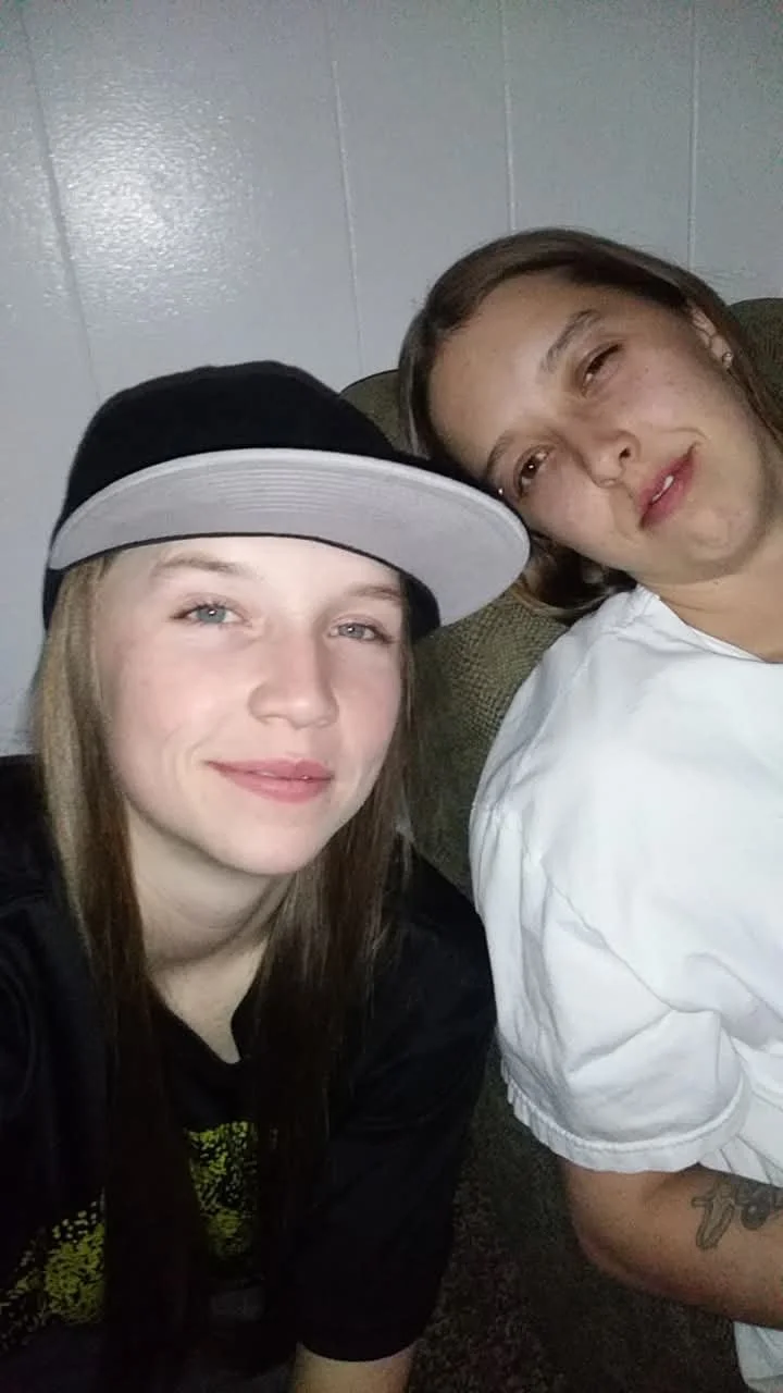 Two young women taking a selfie together, sitting on a couch in a room with white walls. One woman is wearing a black cap and a black shirt, and the other woman is wearing a white shirt.