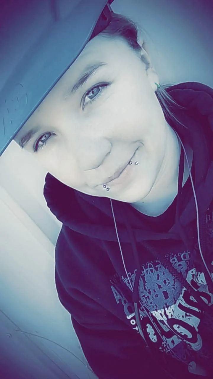 A young person with piercing on their lower lip and a septum piercing, wearing a hoodie with a graphic design, taking a selfie while resting their head against a tan surface, possibly a table or armrest.