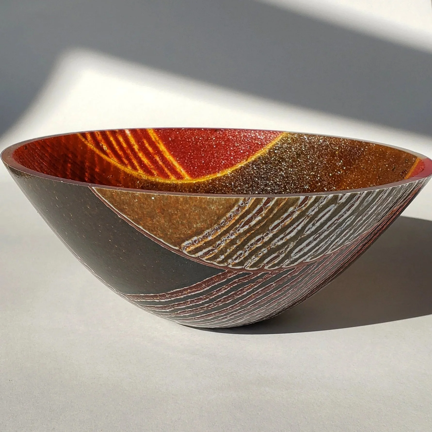 A decorative glass bowl with red, orange, and brown swirling patterns, placed on a white surface with shadow and light.