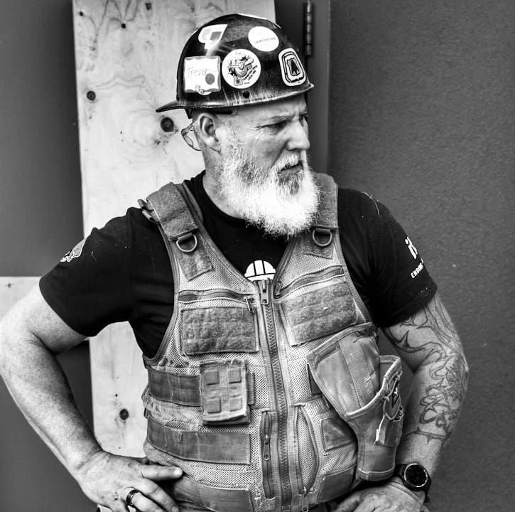 An older man with a beard wearing a safety helmet decorated with stickers, a black T-shirt, a tactical vest, and a wristwatch, standing outdoors against a wall.