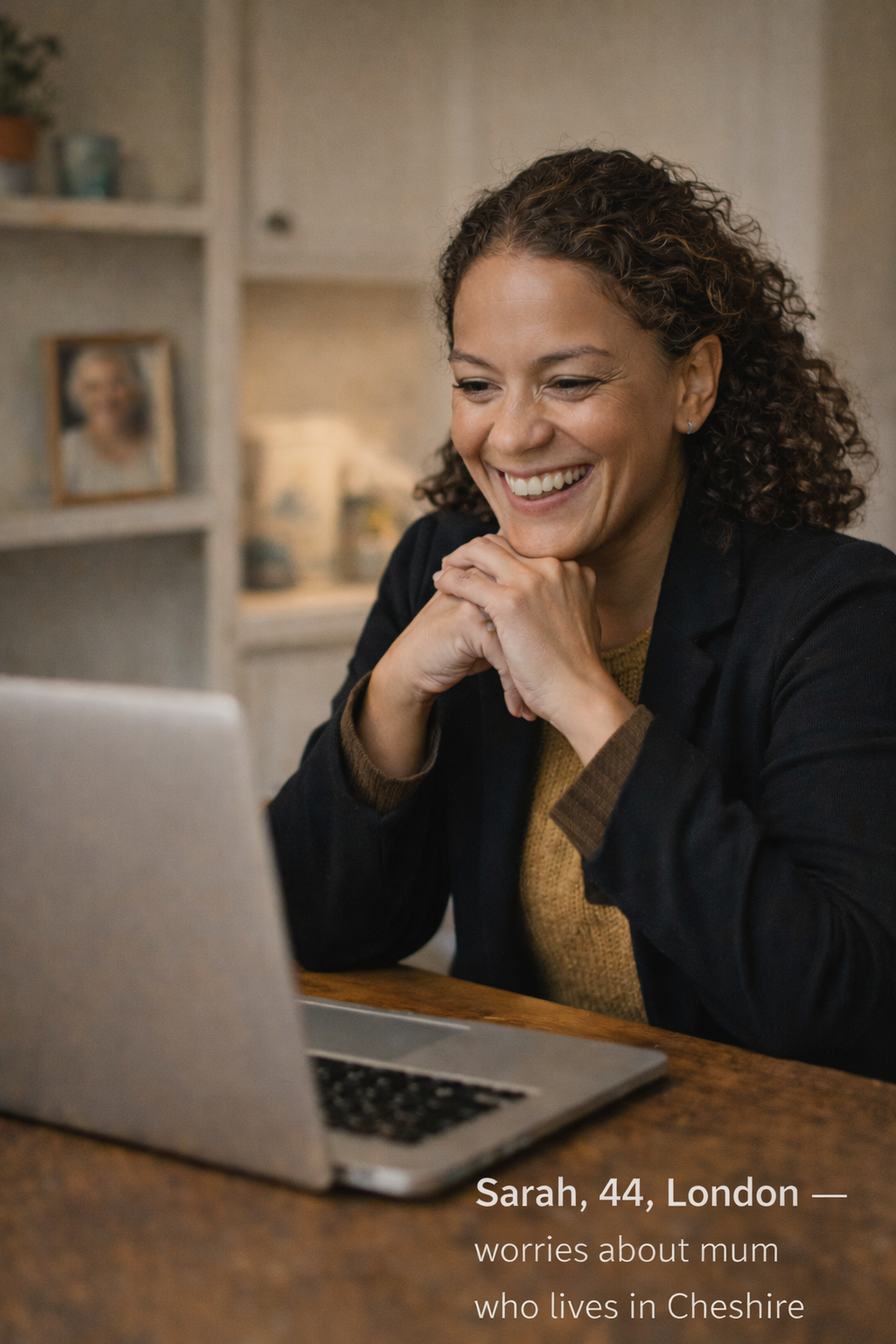 A woman with curly hair smiling and looking at her laptop screen, sitting at a wooden table in a cozy, well-lit room. There are shelves with decorative items and a framed photo in the background. The caption reads: Sarah, 44, London — worries about mum who lives in Cheshire.