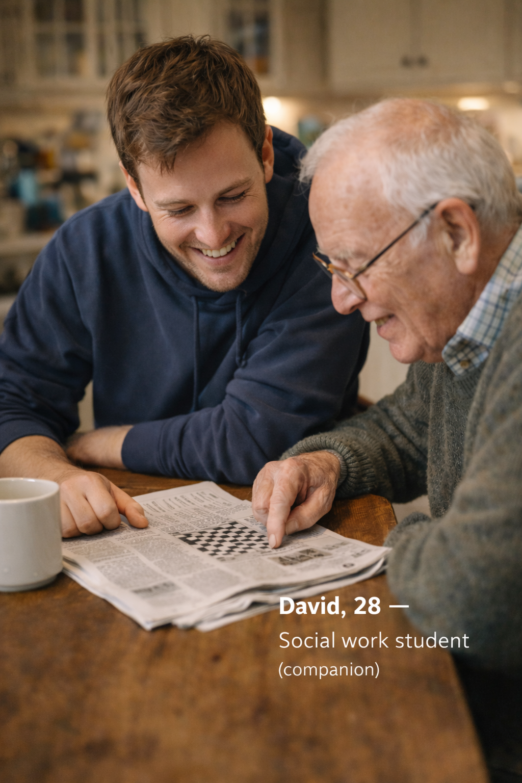 A young man and an elderly man are sitting at a table, smiling and looking at a newspaper or magazine with a chessboard illustration. The young man, identified as David, 28, a social work student and companion, is wearing a navy hoodie. The elderly man is wearing glasses and a gray sweater. There is a white mug on the table.
