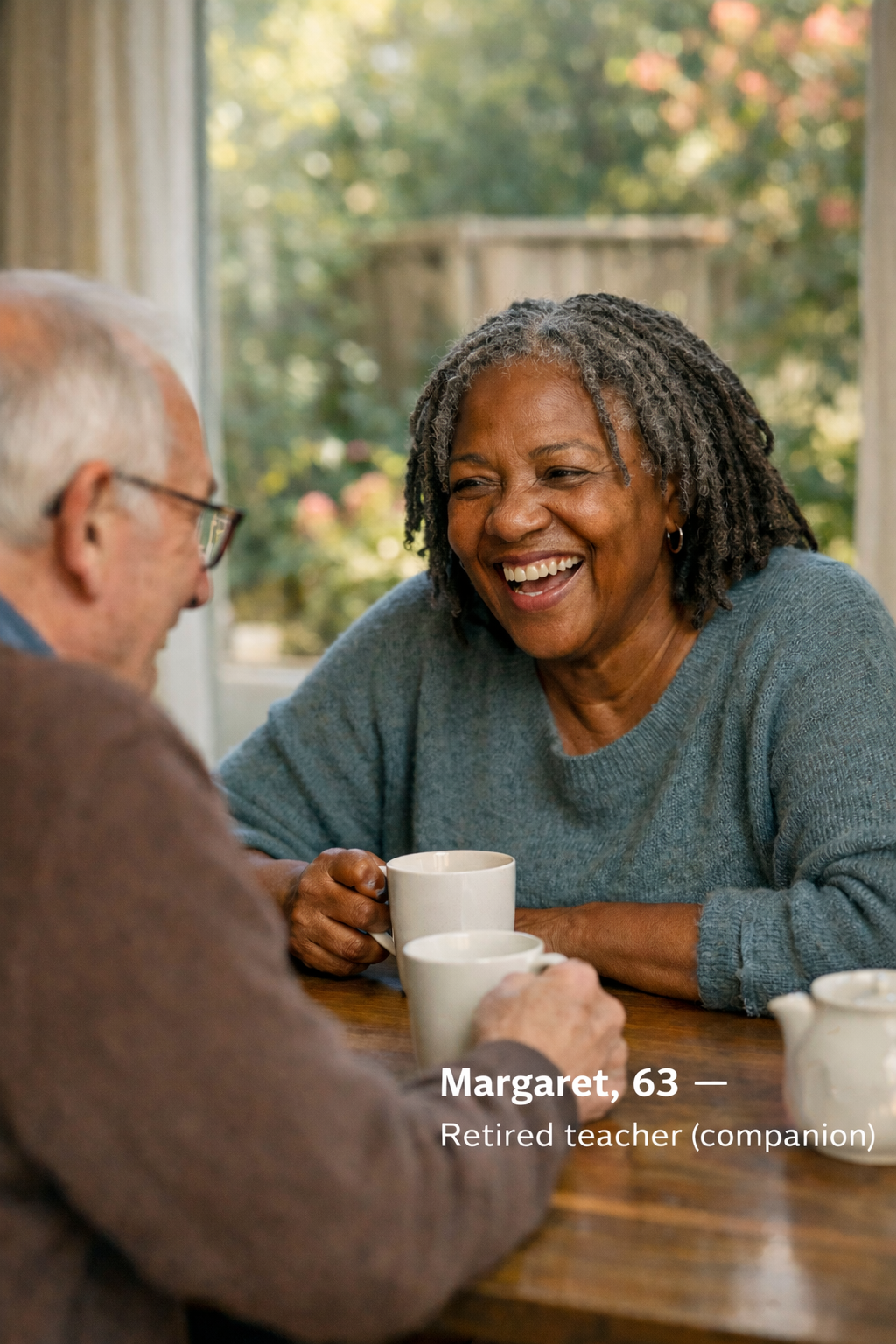 A retired teacher named Margaret, 63, with gray dreadlocks and a blue sweater, smiling and talking with a man with glasses, wearing a brown sweater, during a coffee conversation at a wooden table in a well-lit room with a window showing green trees outside.