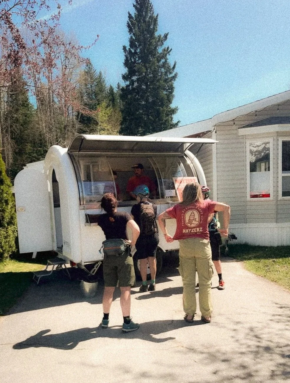 People at a food truck or ice cream stand outdoors near a white building on a sunny day.