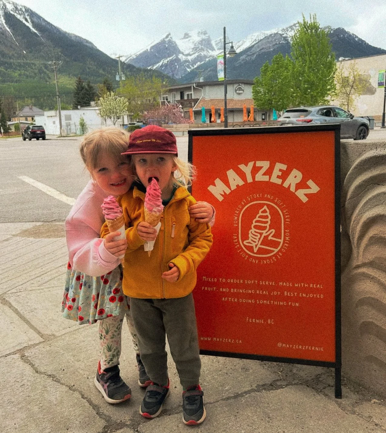 Two young children, a girl and a boy, happily holding ice cream cones in front of a bright red Mayzerz ice cream sign on a sidewalk. The girl is smiling and the boy is licking his ice cream. Behind them, a mountainous landscape with snow-capped peaks and a partly cloudy sky is visible.