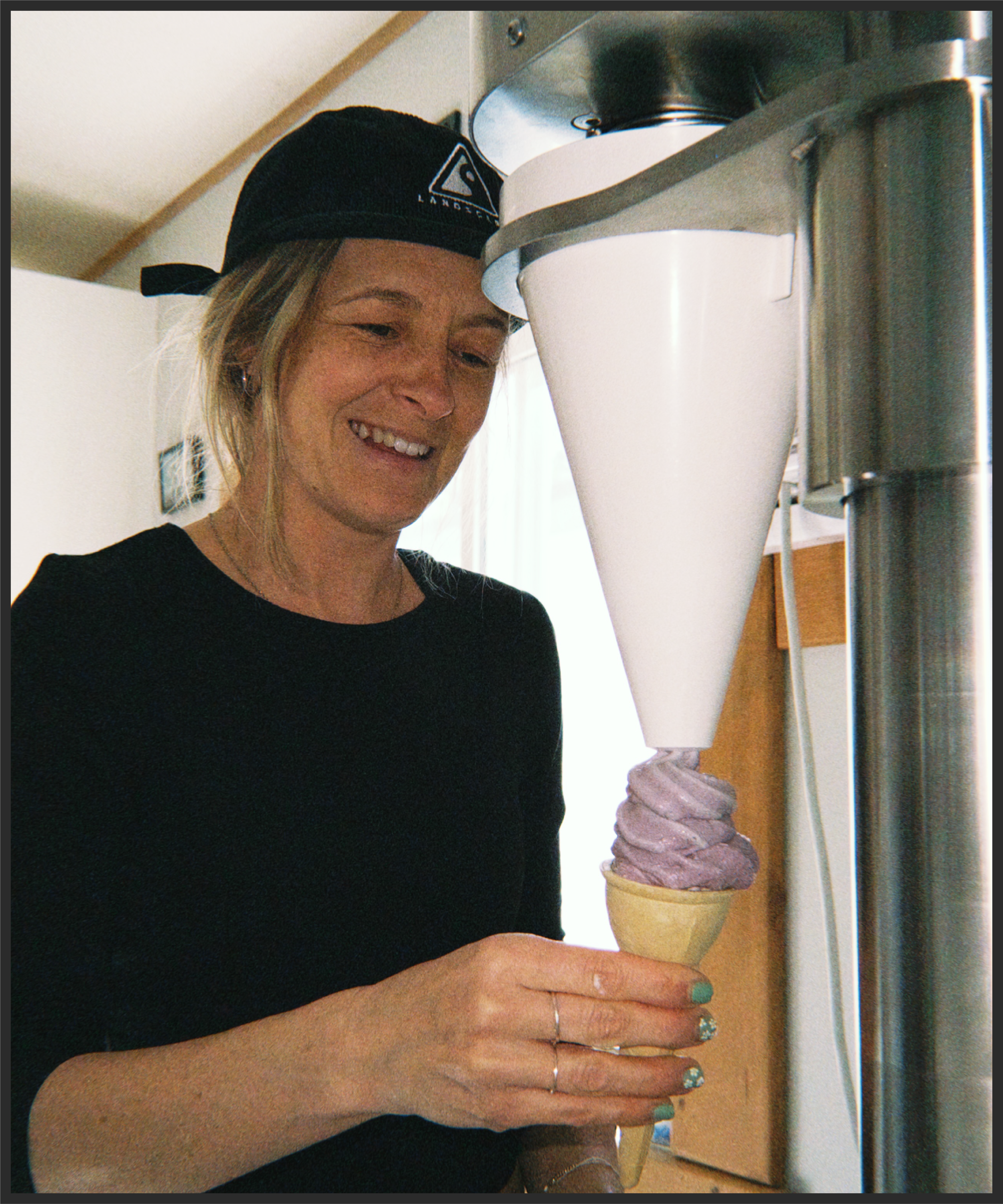 A person with blonde hair, wearing a black beanie with a logo, and a black shirt, is holding a vanilla ice cream cone with a chocolate swirl on top.