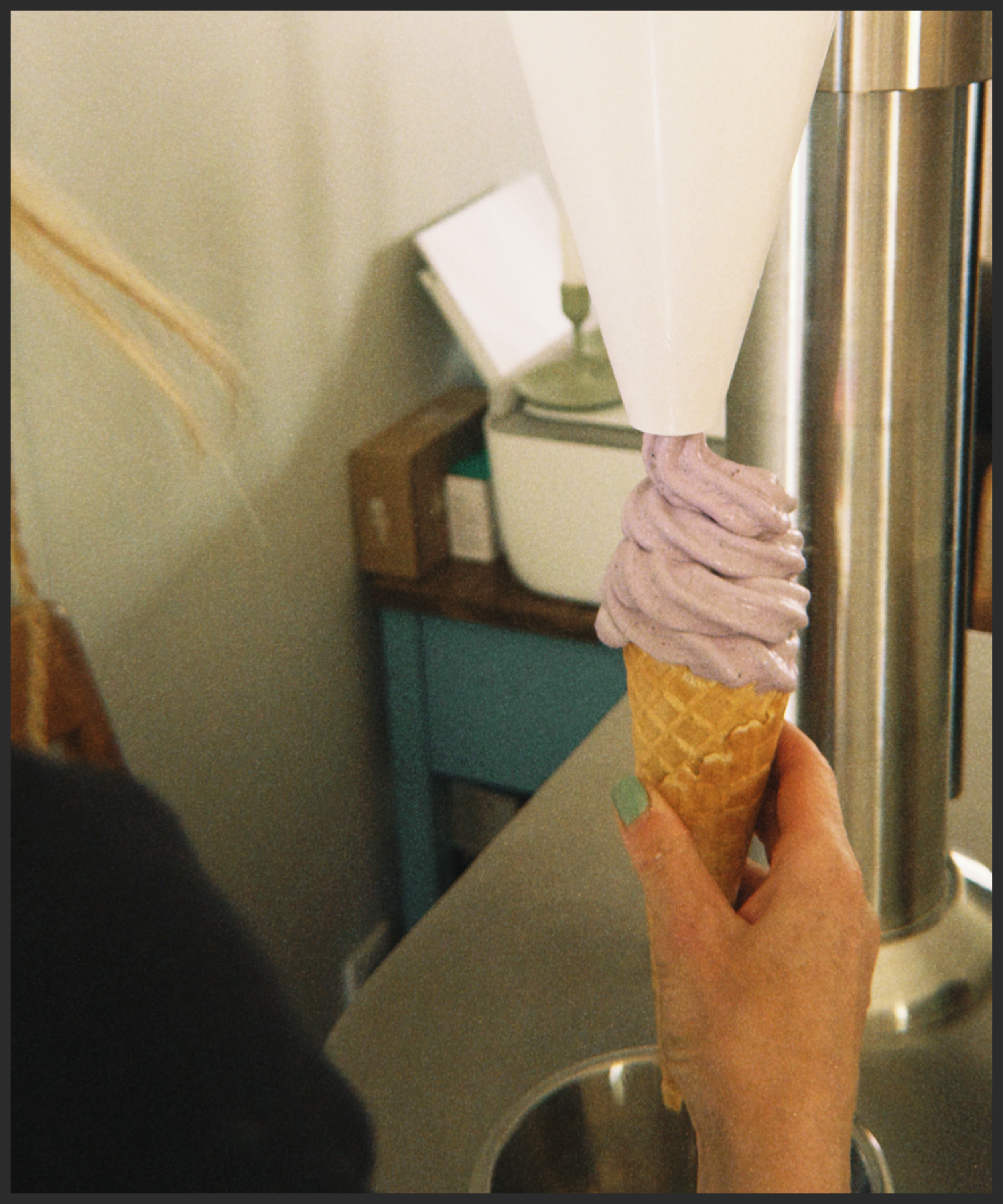 A person holding a soft-serve ice cream cone with pink frosting and sprinkles, standing next to a stainless steel refrigerator in a kitchen setting.