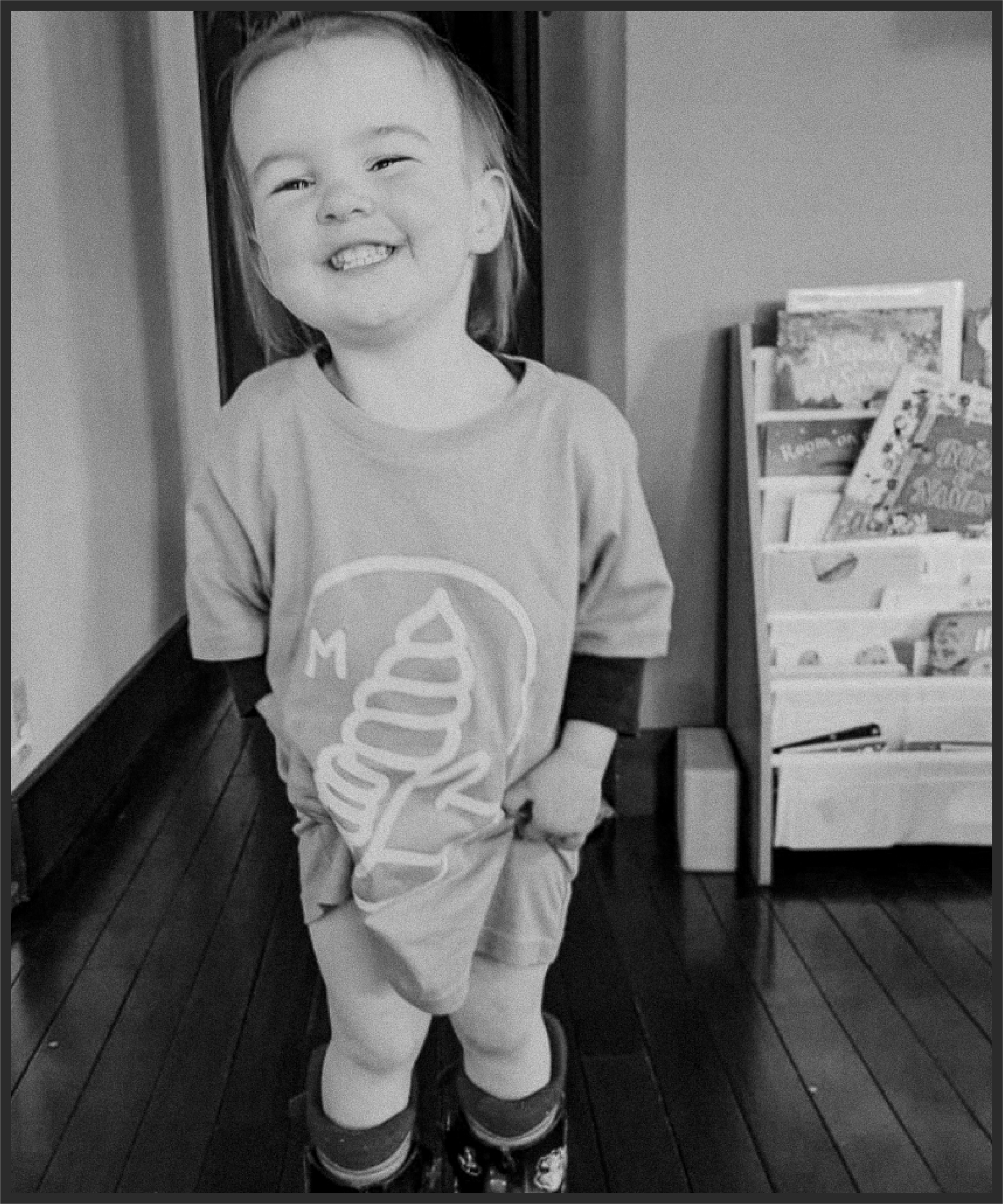 A young child standing indoors, smiling and showing missing teeth, wearing a T-shirt with a graphic design, shorts, knee-high socks, and sneakers, with a bookshelf filled with books in the background.