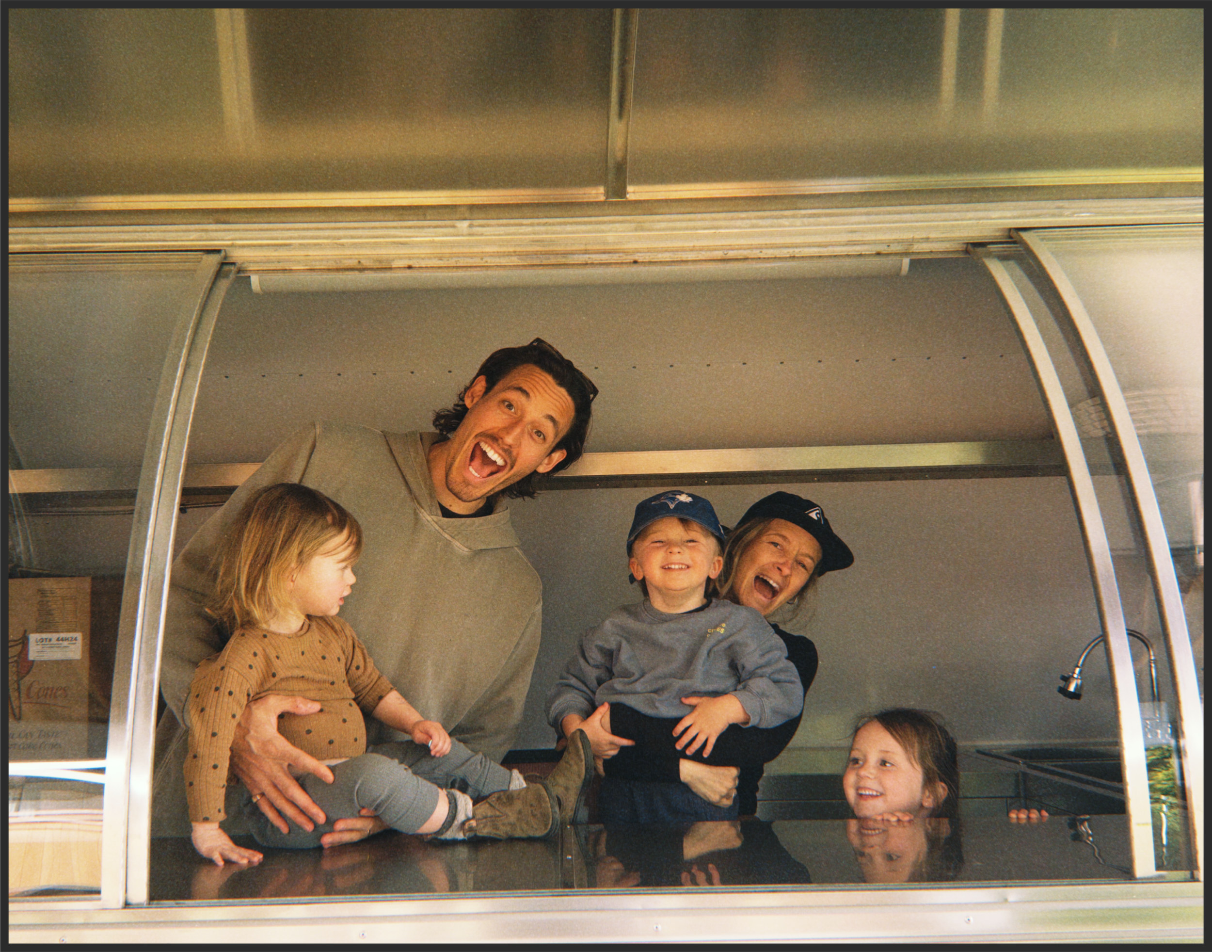 A group of people, including children and adults, are smiling and posing for a photo inside a glass structure with a stainless steel roof and a faucet visible on the right side.