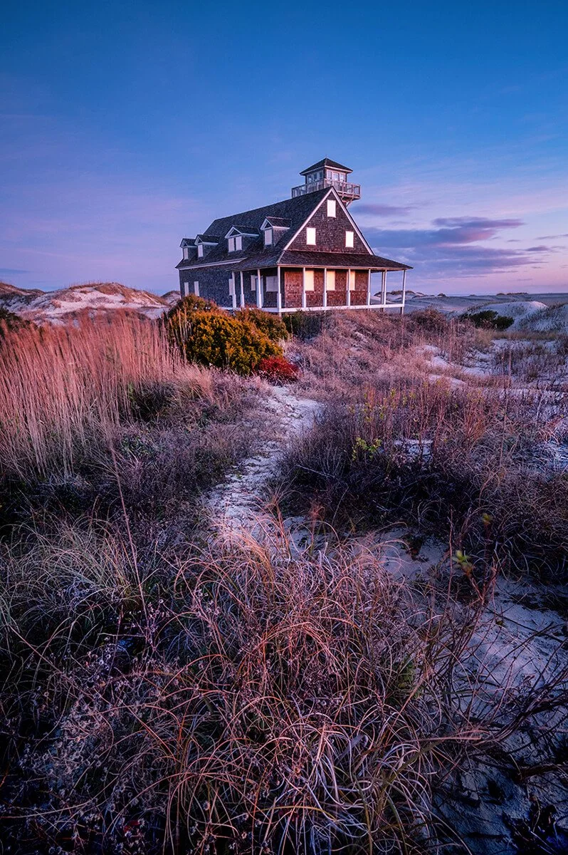 Oregon Inlet Life-Saving Station at daybreak during winter, with coastal grasses in the foreground and a trail leading to the station, under a blue sky with purple overtones. Landscape and coastal photography highlighting historic architecture, seren