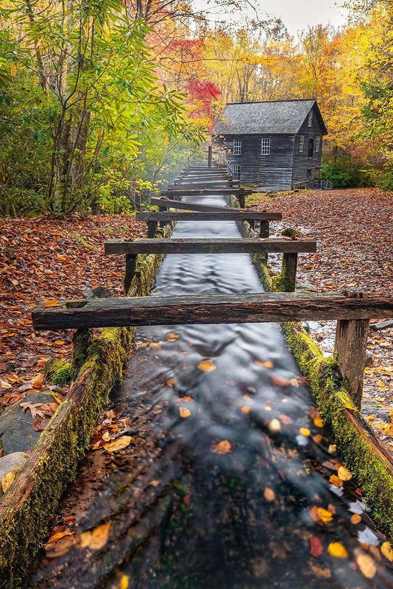 Wide-angle view of the wooden flume at Mingus Mill in the Great Smoky Mountains National Park, with motion-blurred water in the foreground and the flume acting as a leading line to the mill. Vibrant fall colors highlight the scenic landscape and hist