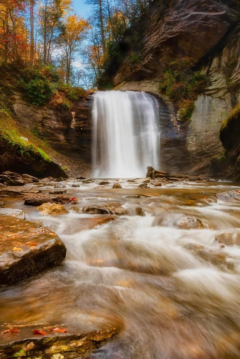 Looking Glass Falls in North Carolina with water flowing over foreground rocks, waterfall in the background under a blue sky.