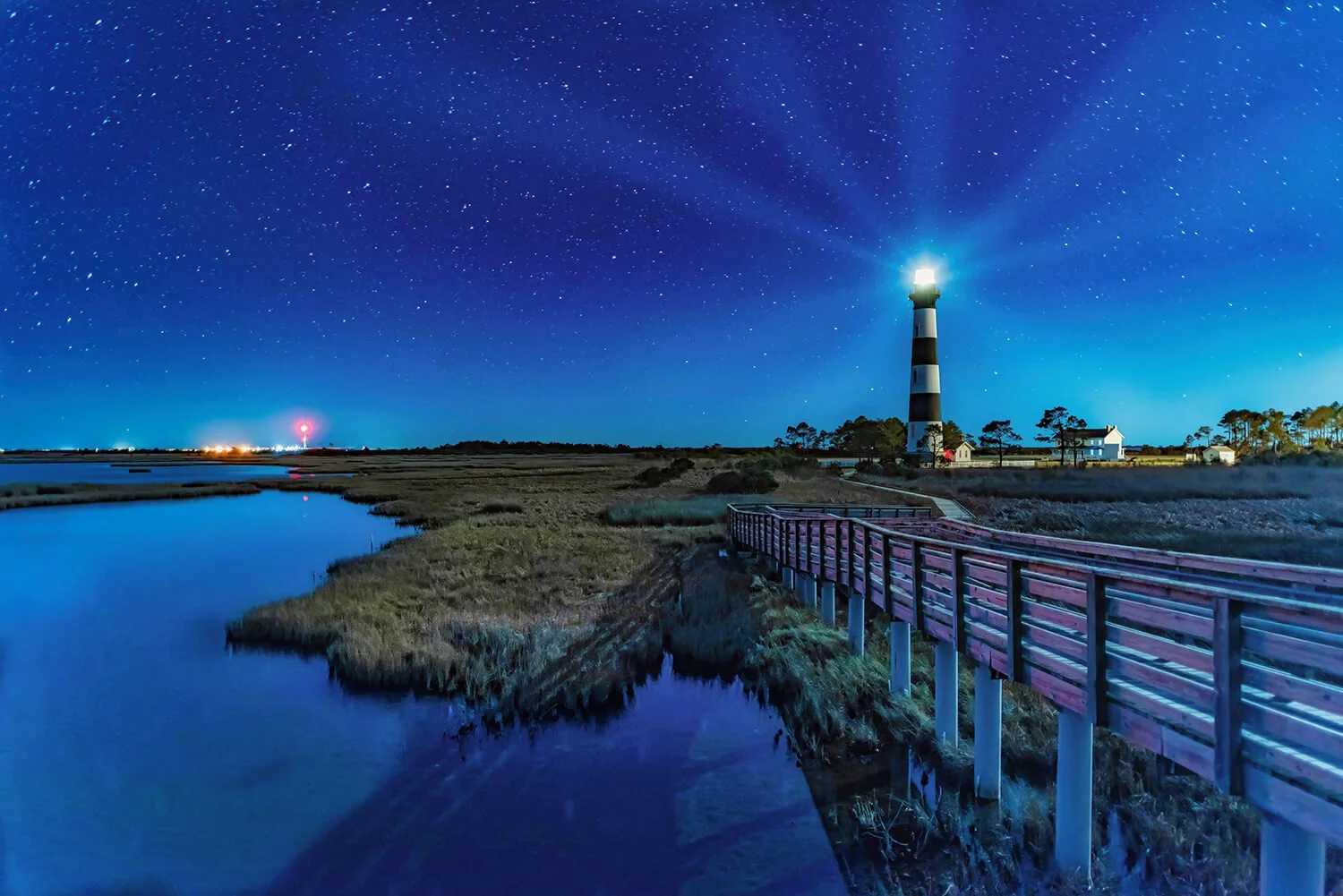 Bodie Island Lighthouse in the Outer Banks of North Carolina at night with stars in a winter-blue sky, lighthouse light casting beams into the sky, and a walkway leading toward the building on the right.