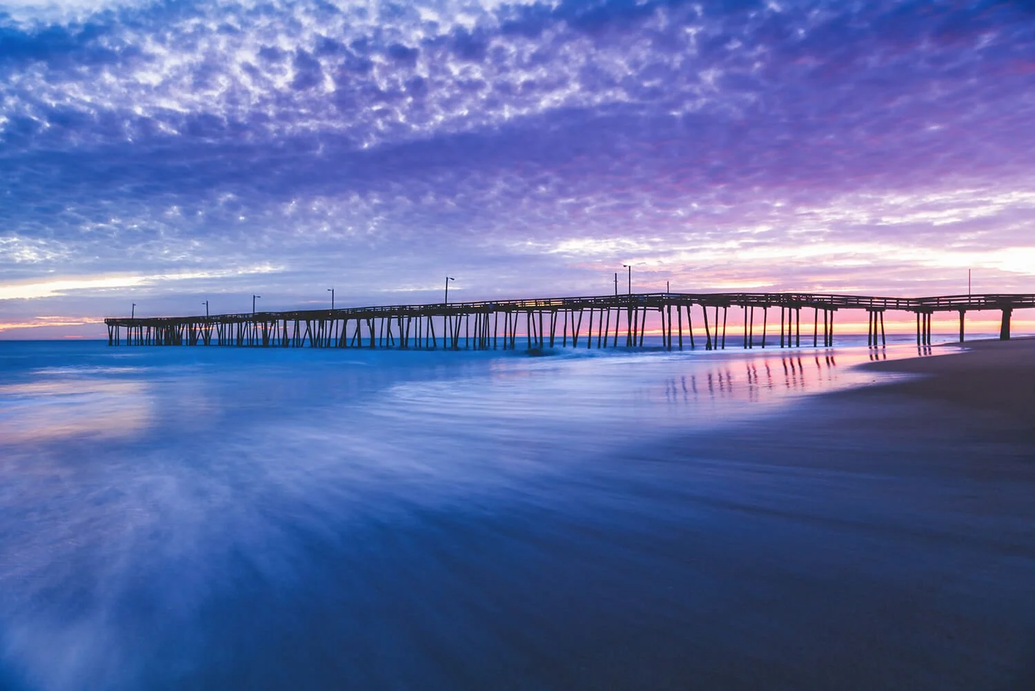 Nags Head Fishing Pier at sunrise with calm receding waters, cloudy skies with deep purple tones, and the pier silhouetted against the rising sun.