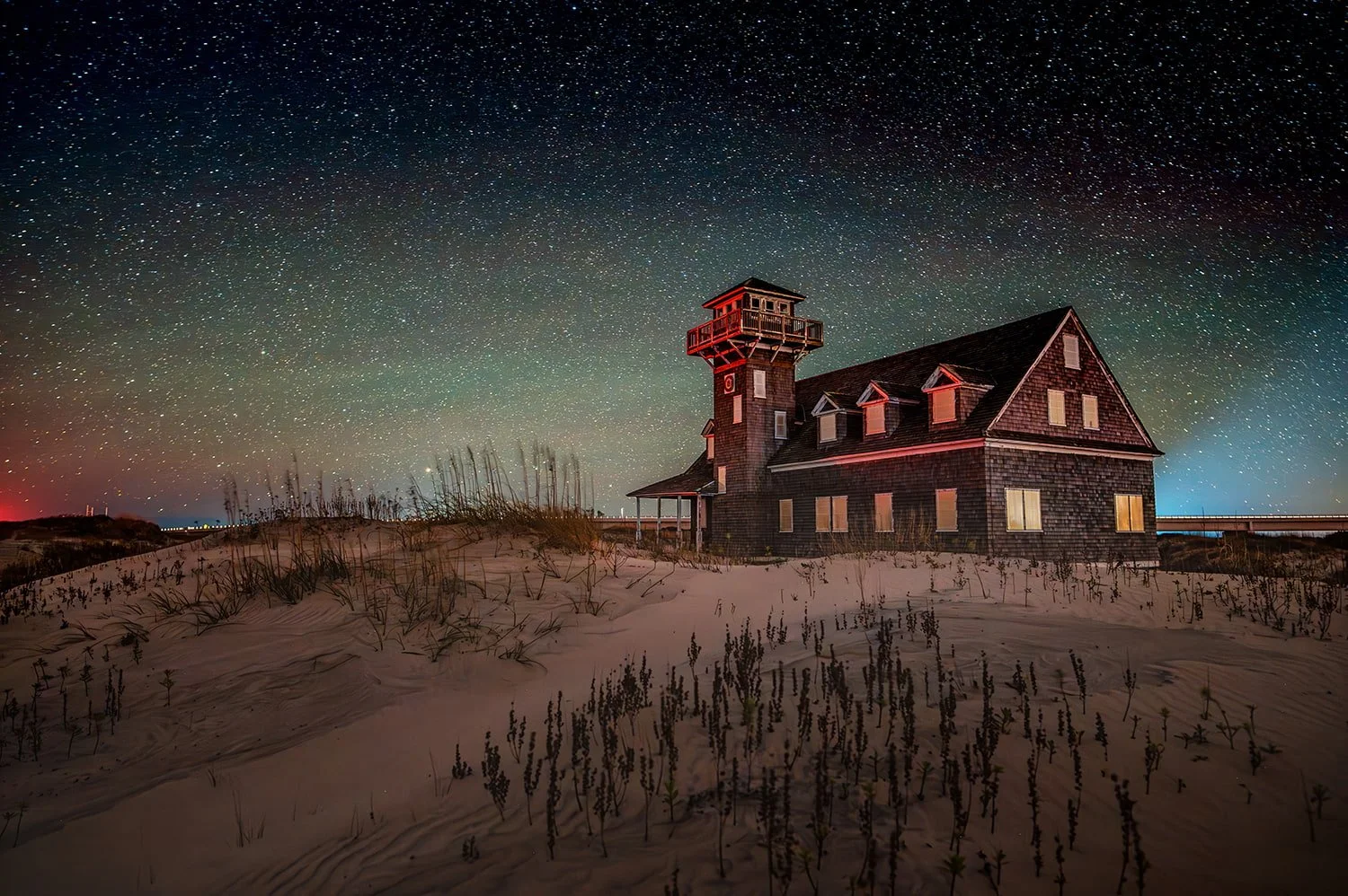 Oregon Inlet Life Saving Station in the Outer Banks of North Carolina at night under a star-filled sky, with a red light in the distance casting a red glow on the tower and windows.