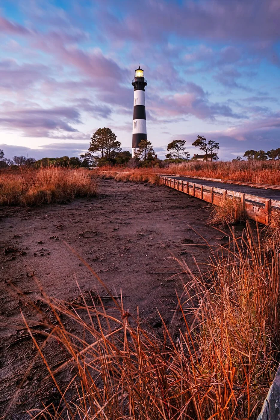 Bodie Island Lighthouse just after sunrise in winter with coastal grass in the foreground, a dry marsh bed in the middle, a deck walkway leading to the lighthouse, its light glowing, and purple-gray clouds moving against a blue sky.