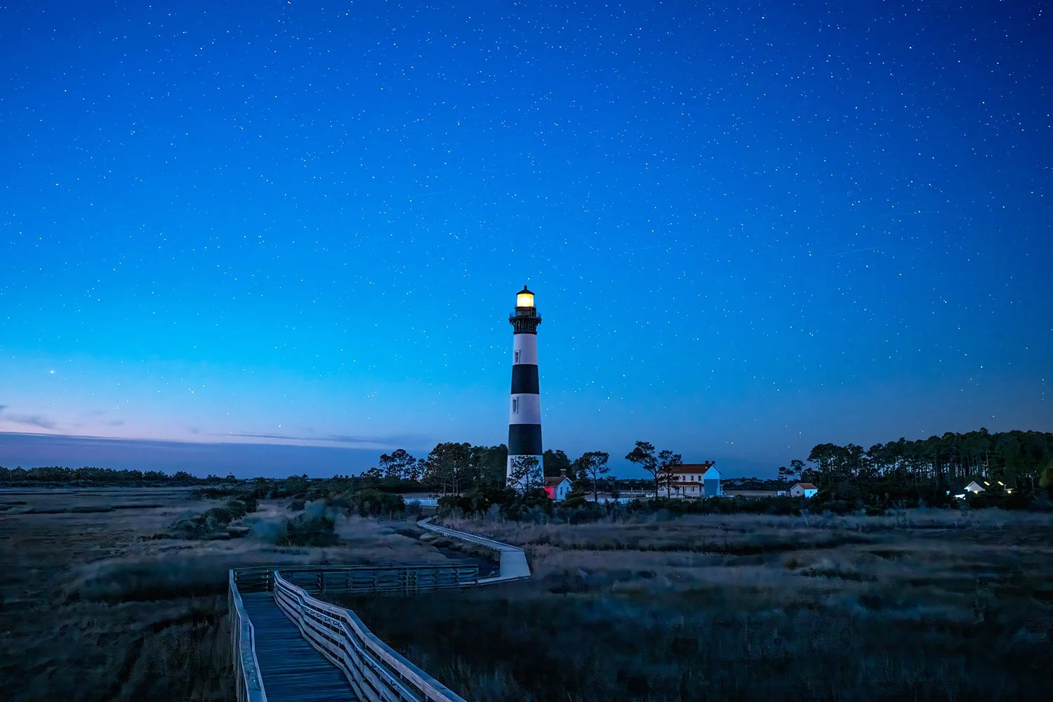 Bodie Island Lighthouse viewed from the observation deck at daybreak, with a deep blue sky dotted with stars and a hint of purple clouds in the lower left.