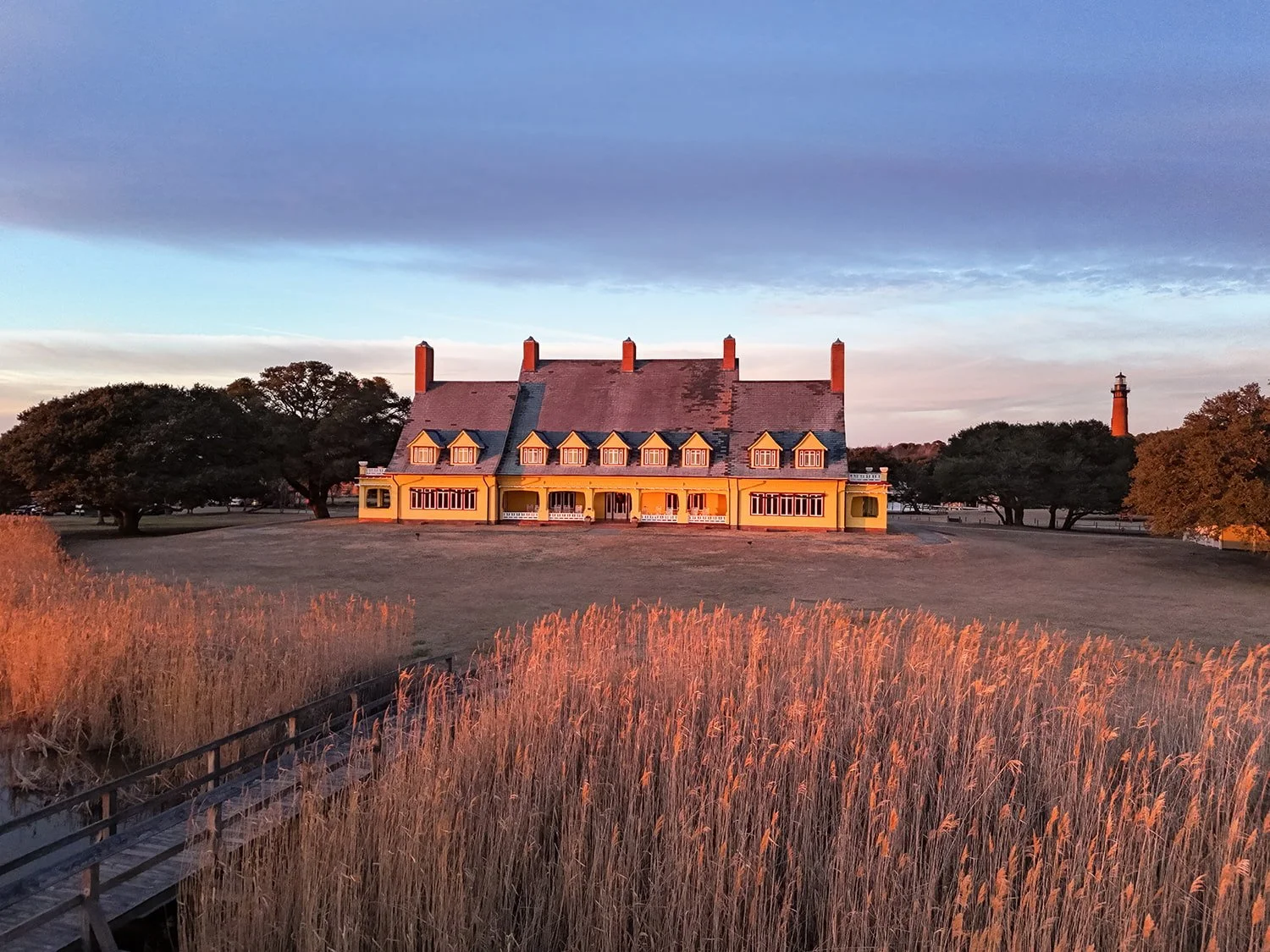 Whale Head Club in Corolla, North Carolina at sunset with the sun illuminating the front of the building and coastal grasses in the pond, a deck walkway leading toward the club, and Currituck Lighthouse visible in the distance to the right.