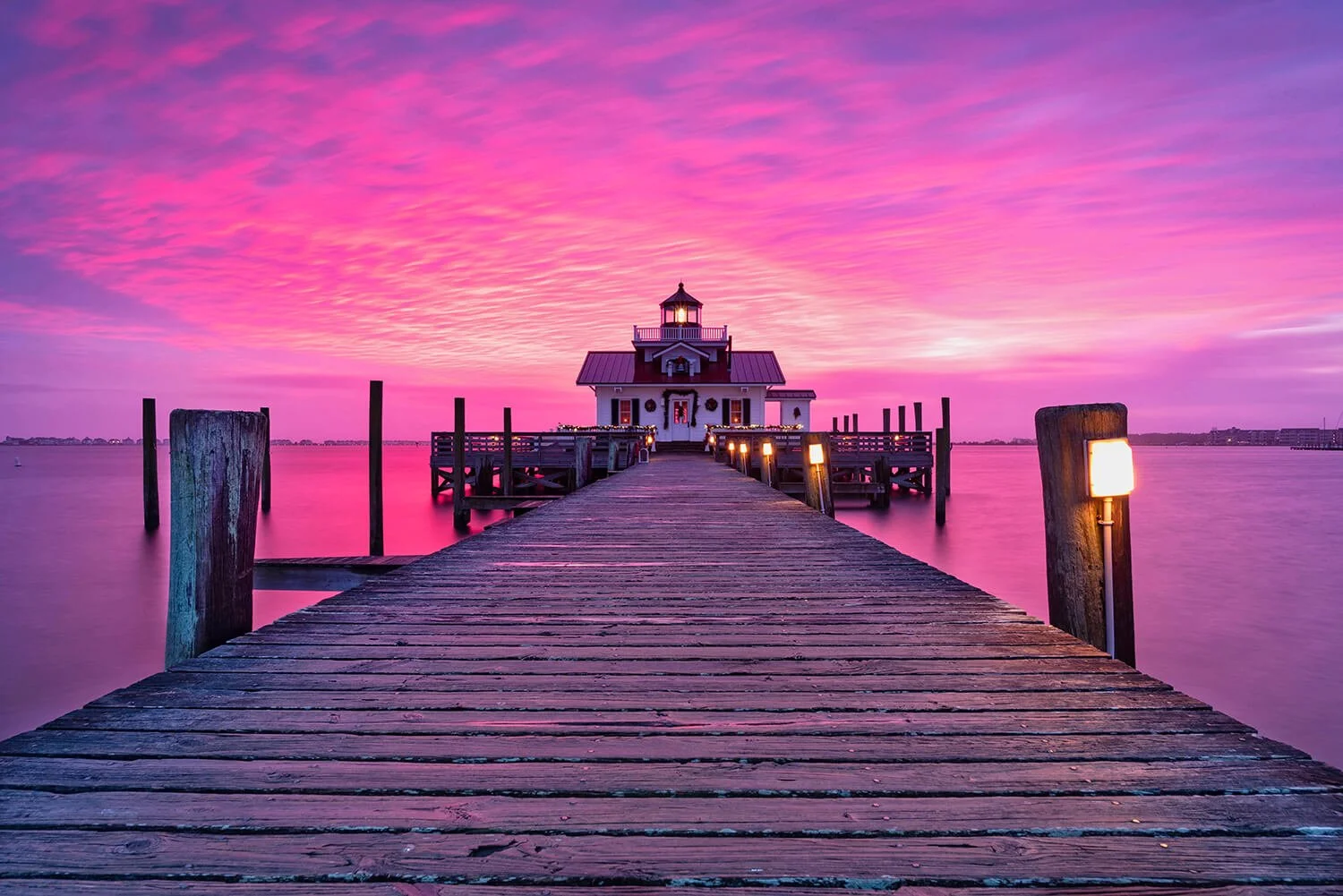 Roanoke Marshes Lighthouse at sunrise with brilliant pink, purple, and orange skies reflecting on the dock, with dock lights illuminated.