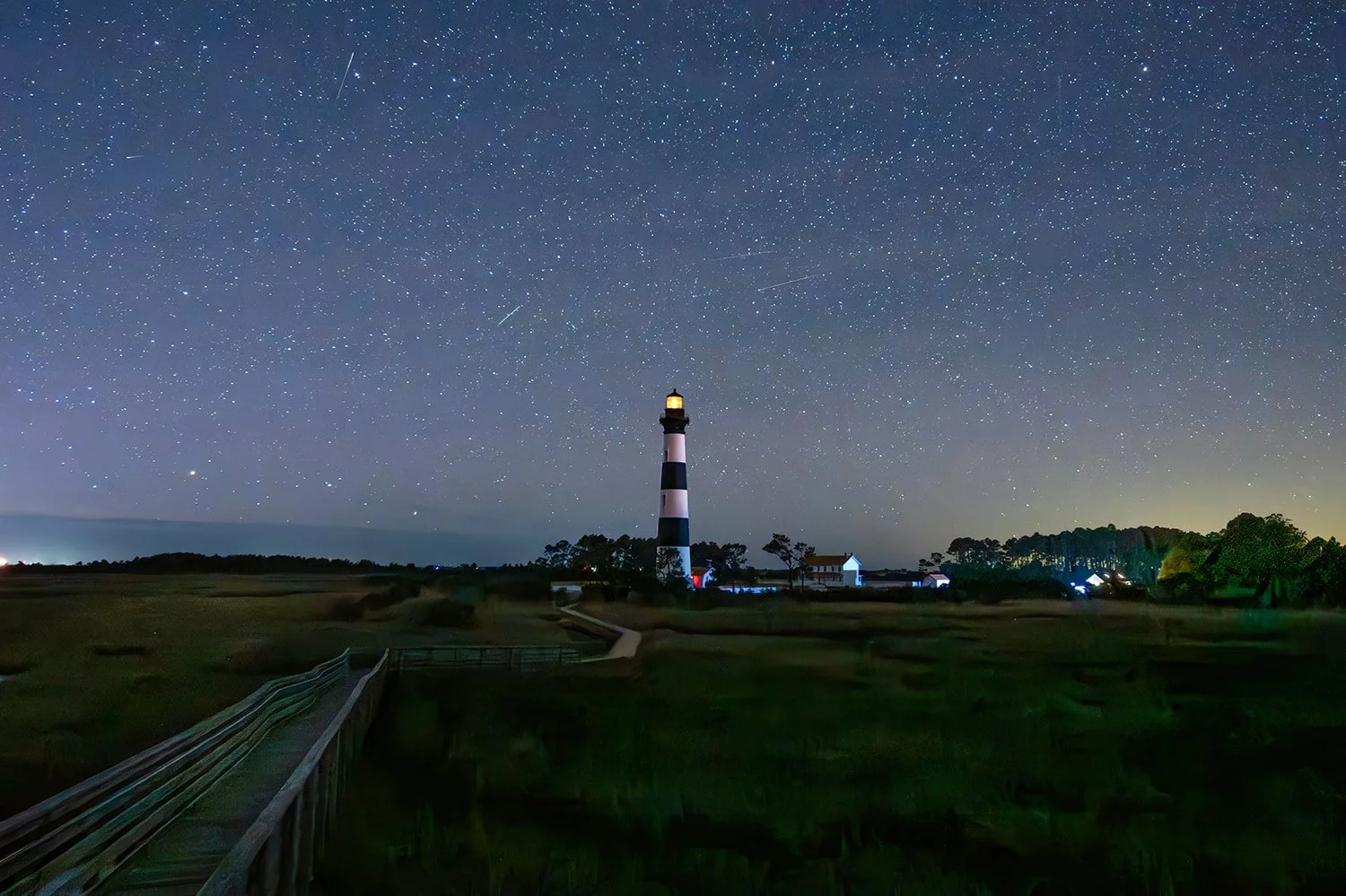 Bodie Island Lighthouse in the Outer Banks of North Carolina at night under a star-filled sky, centered in a wide shot from the observation deck with its light glowing.