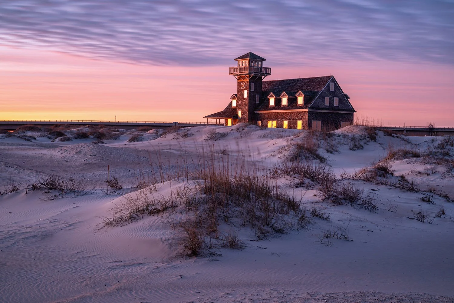 Oregon Inlet Life Saving Station at sunrise with sand dunes and grasses in the foreground, building glowing orange, and purple and orange clouds in a warm-toned sky.
